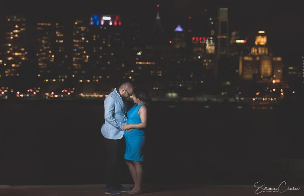 A couple stands close together at night with their foreheads touching, holding hands, in front of a city skyline with illuminated buildings.