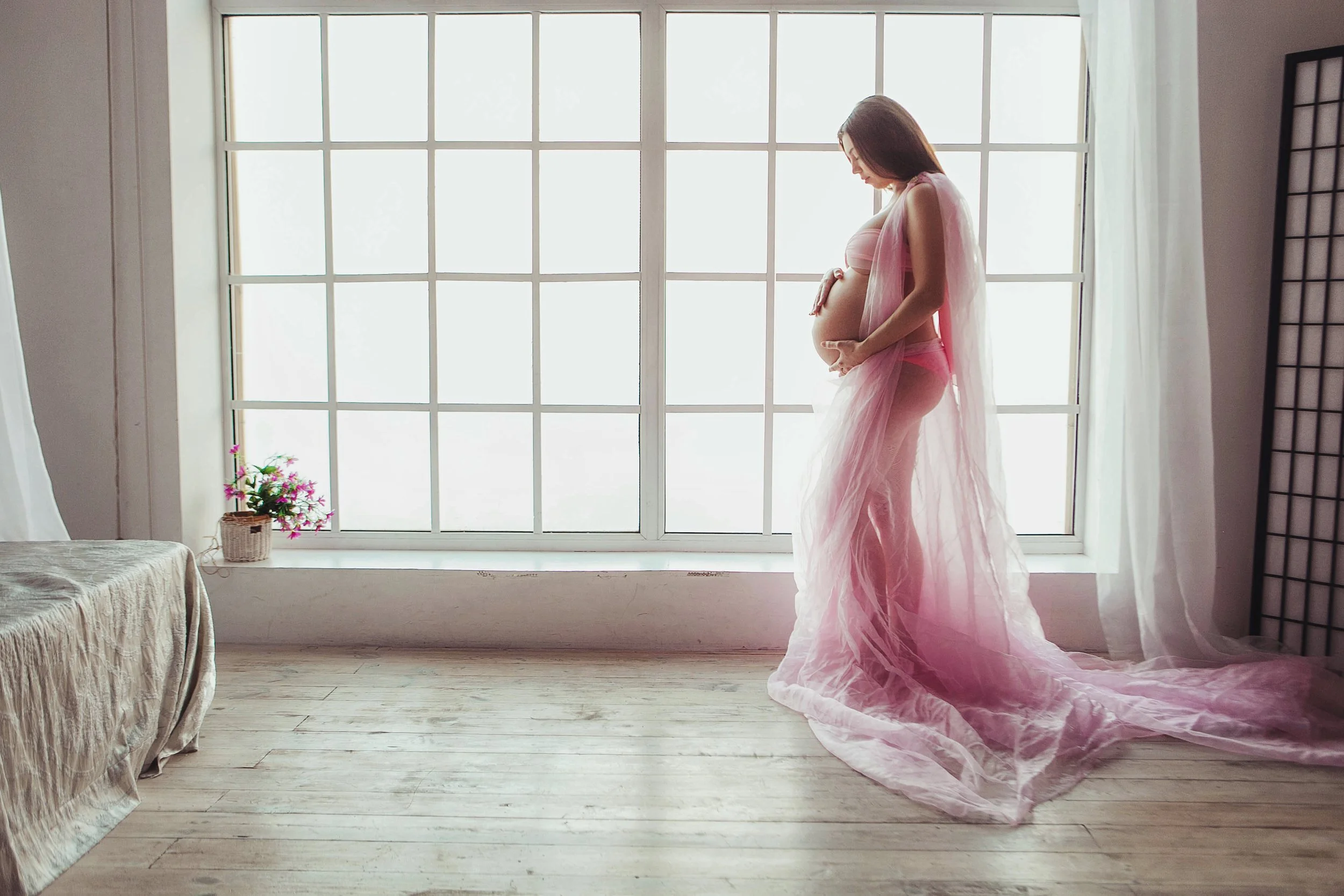 Pregnant woman in pink flowing dress standing by a large window with white curtains, holding her belly.