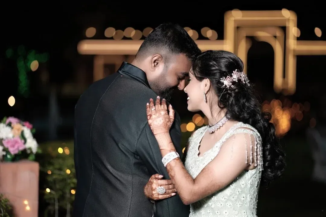 A man proposes to a woman on the beach at sunset under a decorated canopy with the words 'Marry Me' lit up in the background. The man is kneeling with a ring, and the woman appears surprised and emotional.