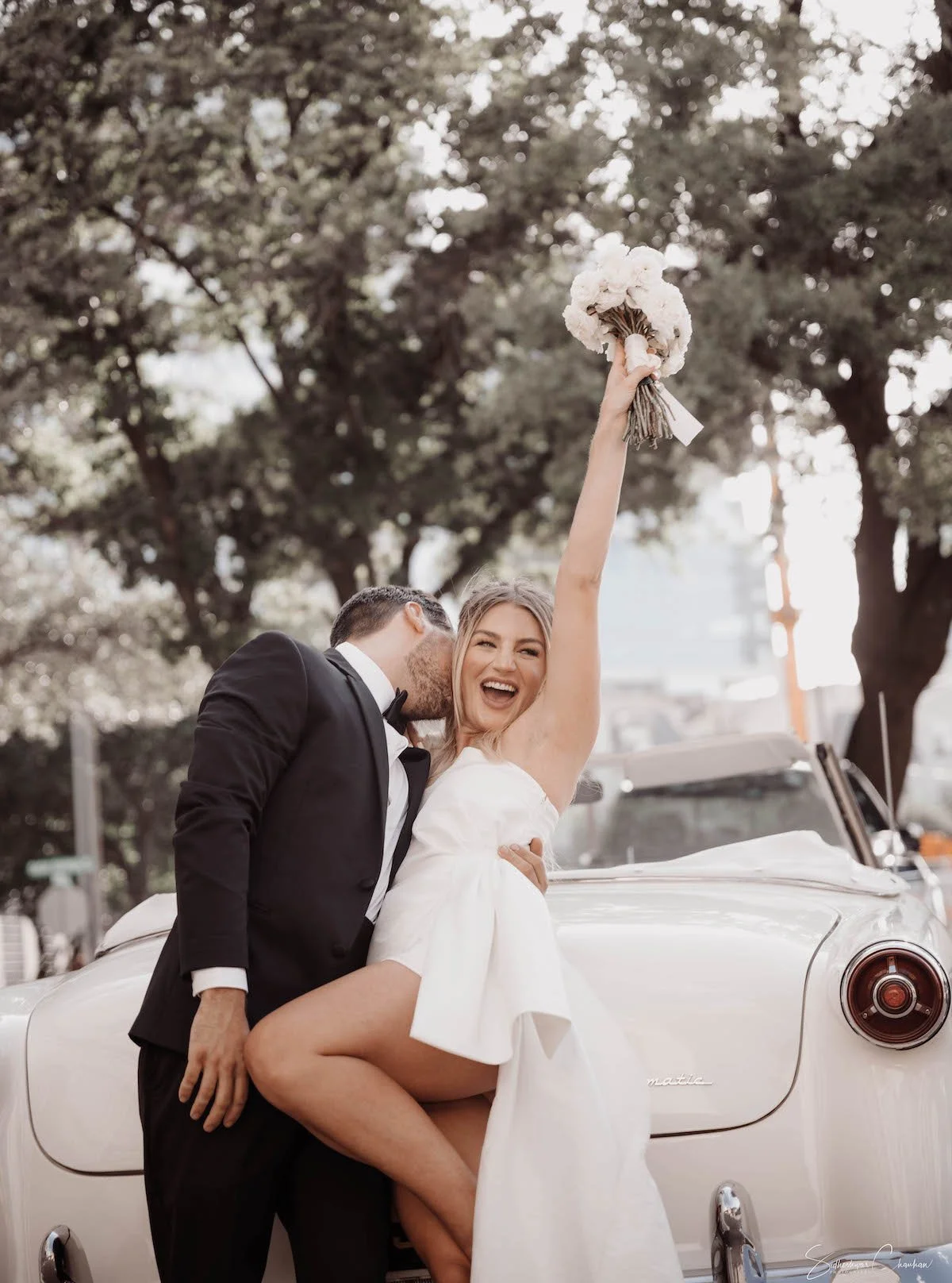 A newlywed couple celebrating in front of a vintage white car, with the bride holding a bouquet of flowers up in the air and the groom embracing her from behind with a kiss on her cheek, outdoors with trees in the background.