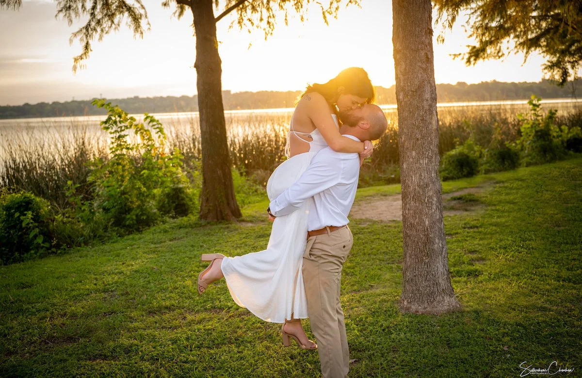 A man in beige pants and a white shirt lifting a woman in a white dress and high heels in the air while kissing in a park at sunset, with trees and a lake in the background.