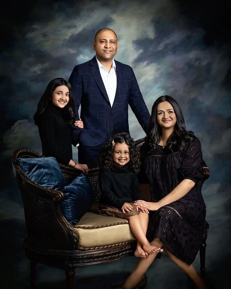 A family portrait of a man, woman, and three young girls posing together in a studio with a dark, cloudy background. The man is standing behind a vintage-style sofa, the woman sitting on it, and the girls are positioned around them.