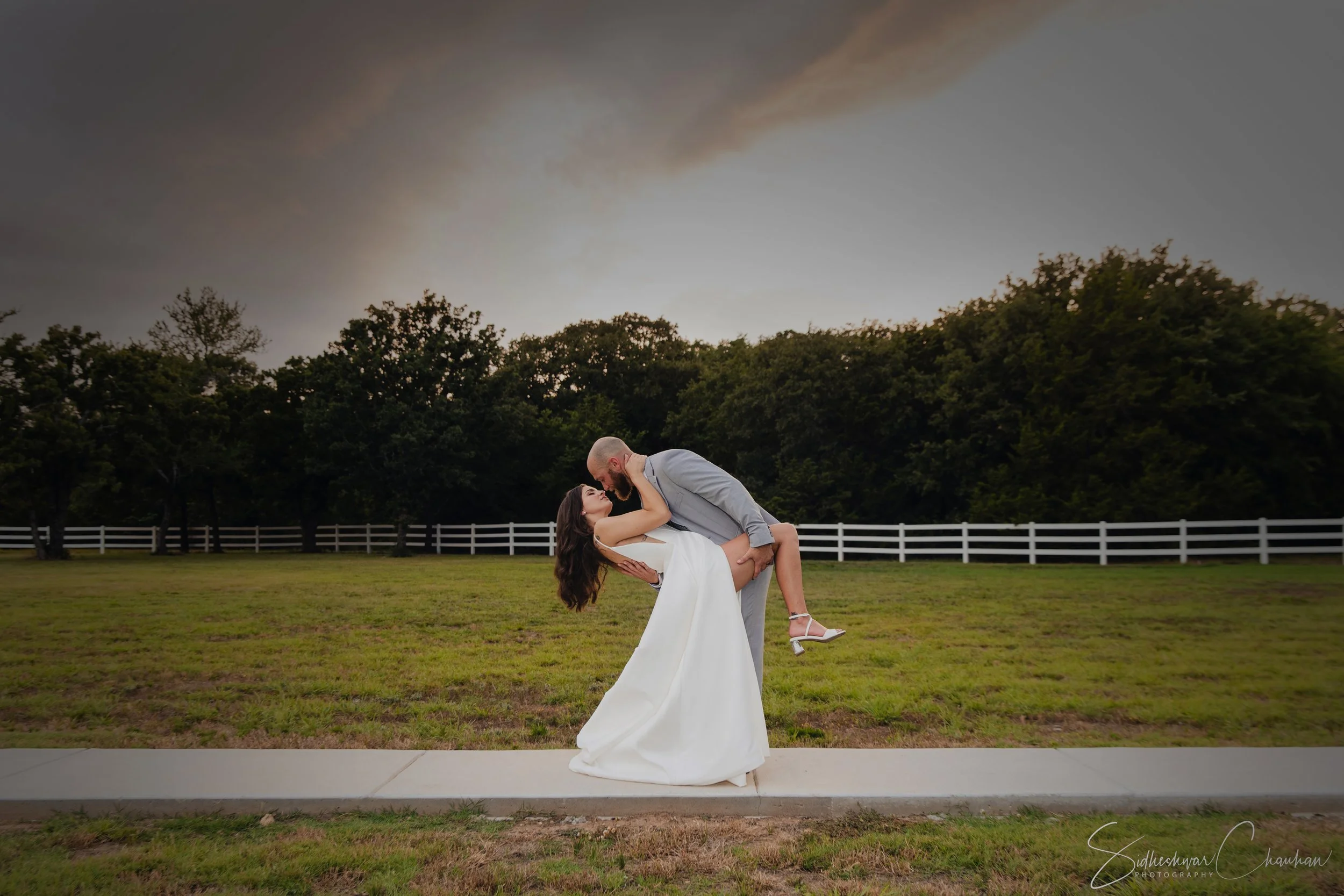 A man and woman in wedding attire share a romantic embrace outdoors; the man, wearing a grey suit, dips the woman in a white wedding dress against a backdrop of trees and darkening sky.