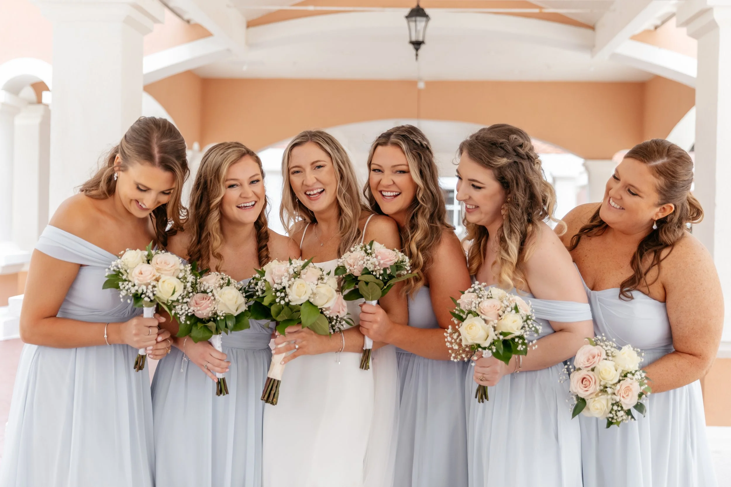 Six women in light blue dresses, with one woman in a white dress, holding bouquets of white and pink roses, standing together and smiling indoors.