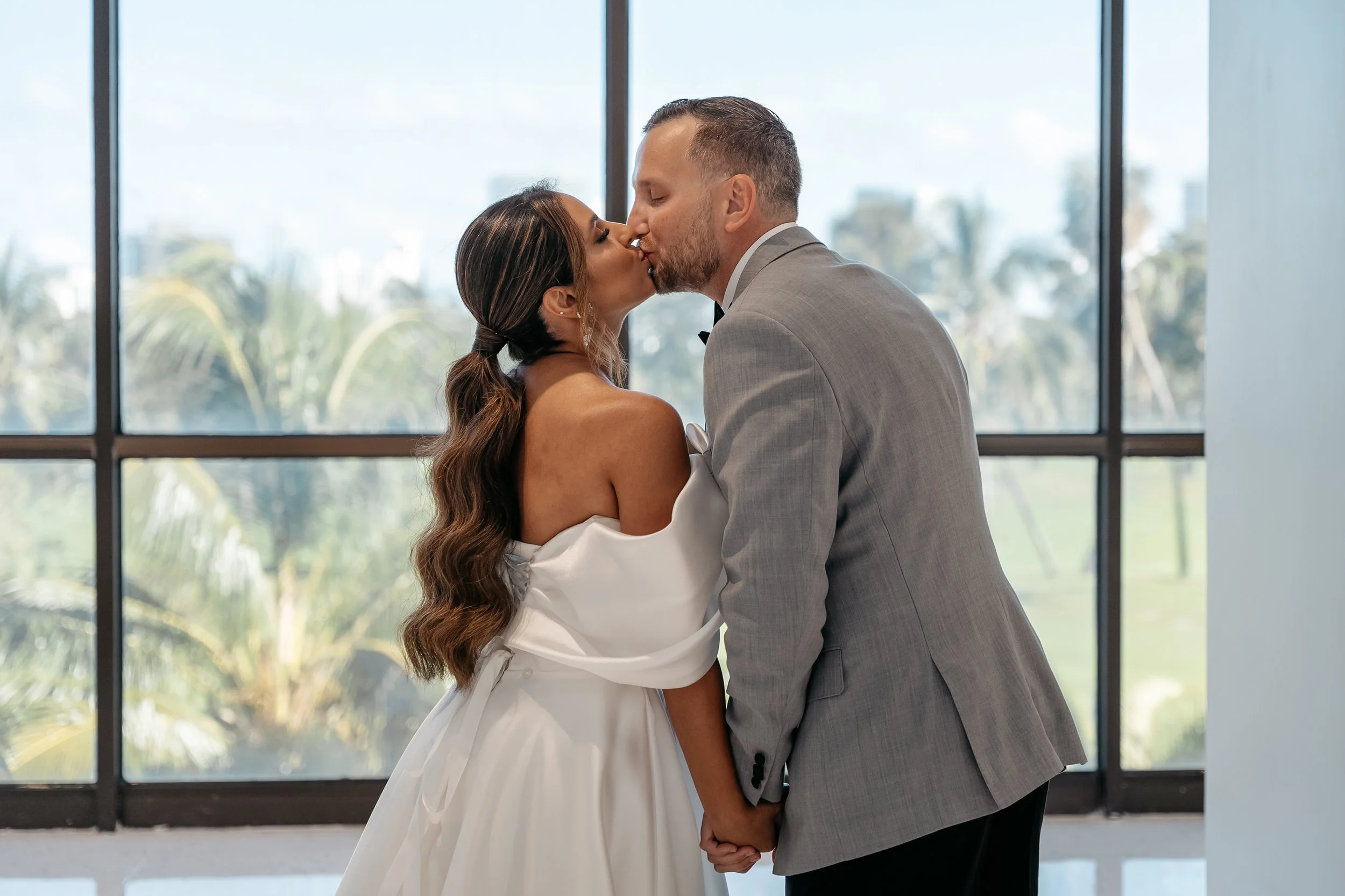 A bride and groom sharing a kiss during their wedding ceremony indoors with large windows showing trees outside.