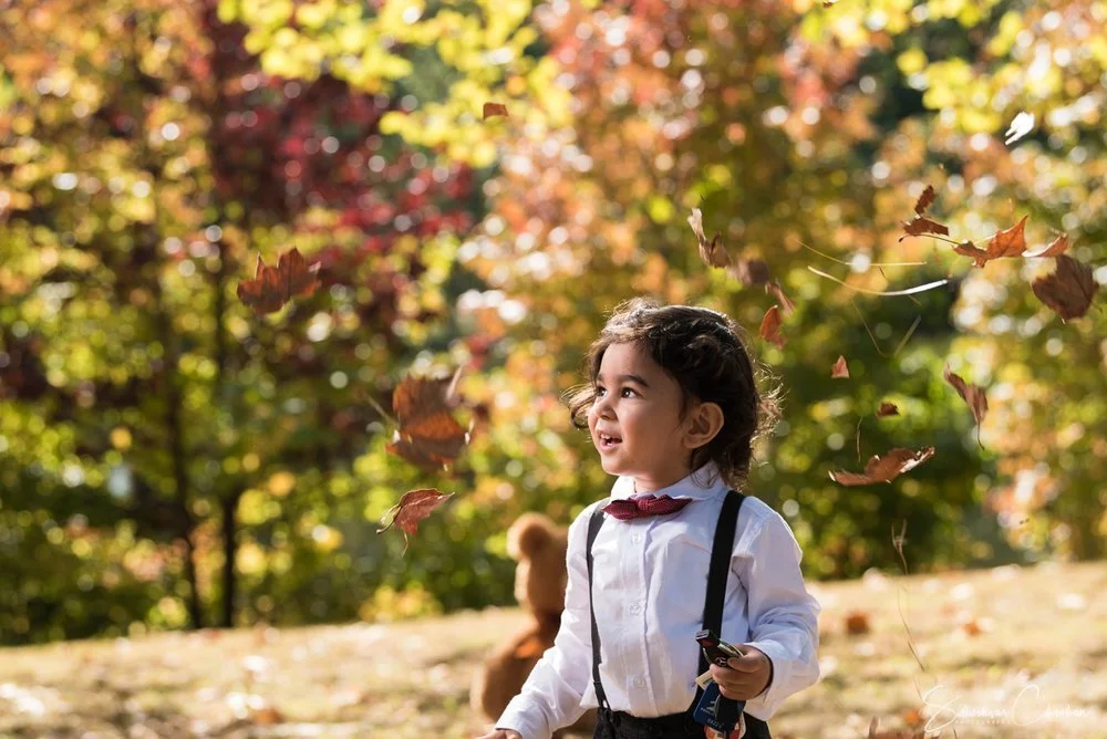 A young girl in a white shirt, black suspenders, and a red bow tie playing outside among falling autumn leaves with colorful trees in the background.