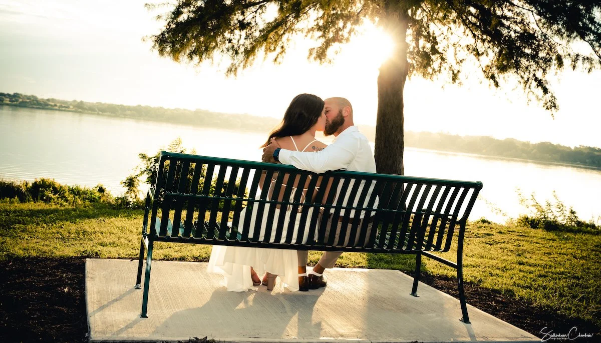 A couple sitting on a park bench by a lake, kissing, with the sun setting behind a tree.