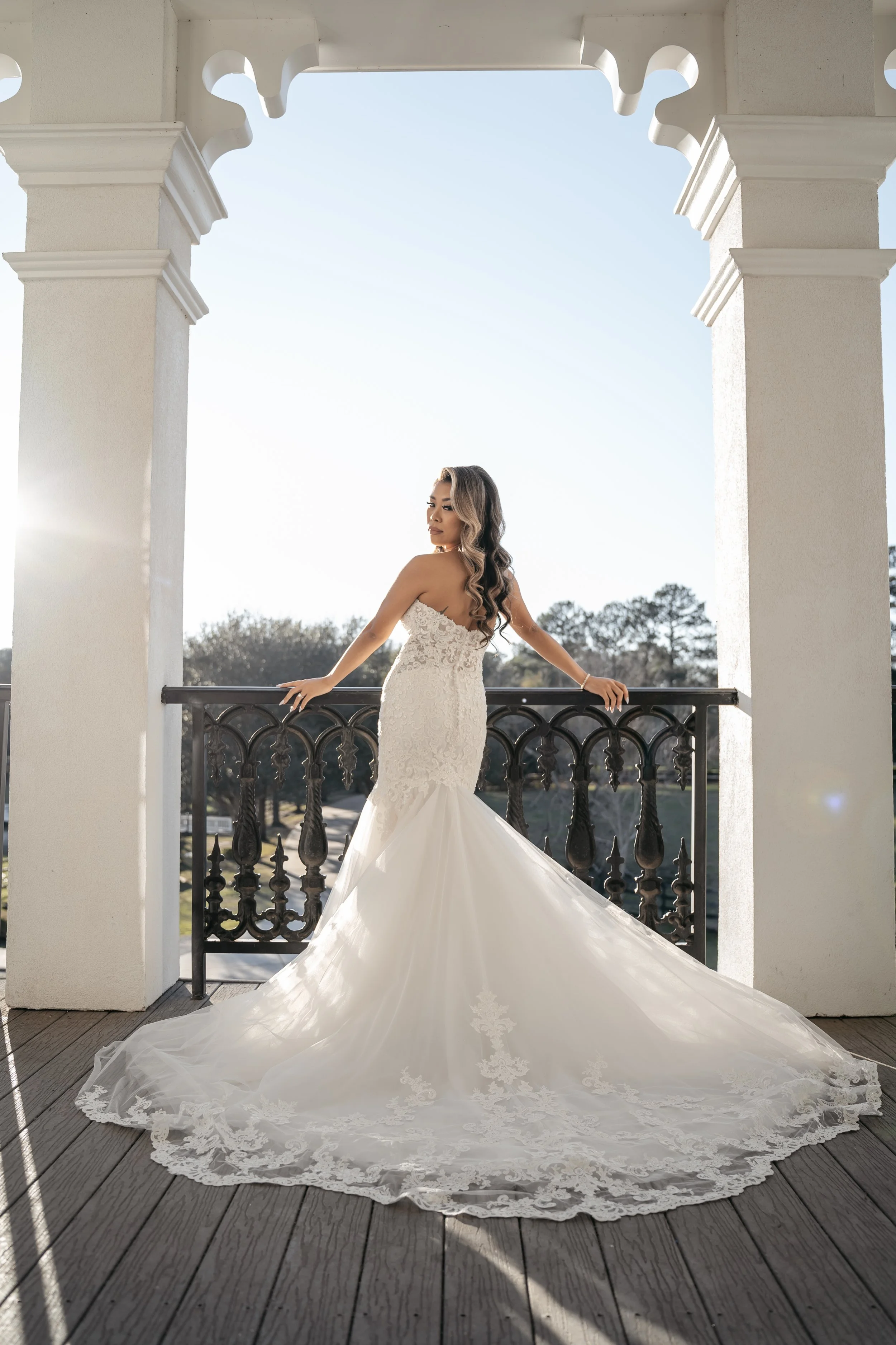 A woman in a wedding dress standing on a balcony with ornate railings, with the sun setting behind her.