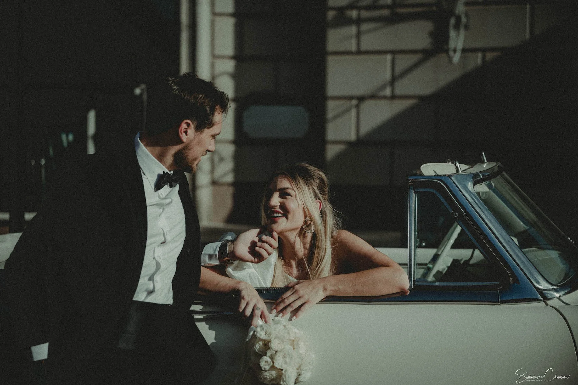 A couple dressed in wedding attire, the man in a tuxedo and the woman in a wedding dress, sharing a joyful moment leaning on a vintage convertible car, with the woman holding a bouquet of white flowers.