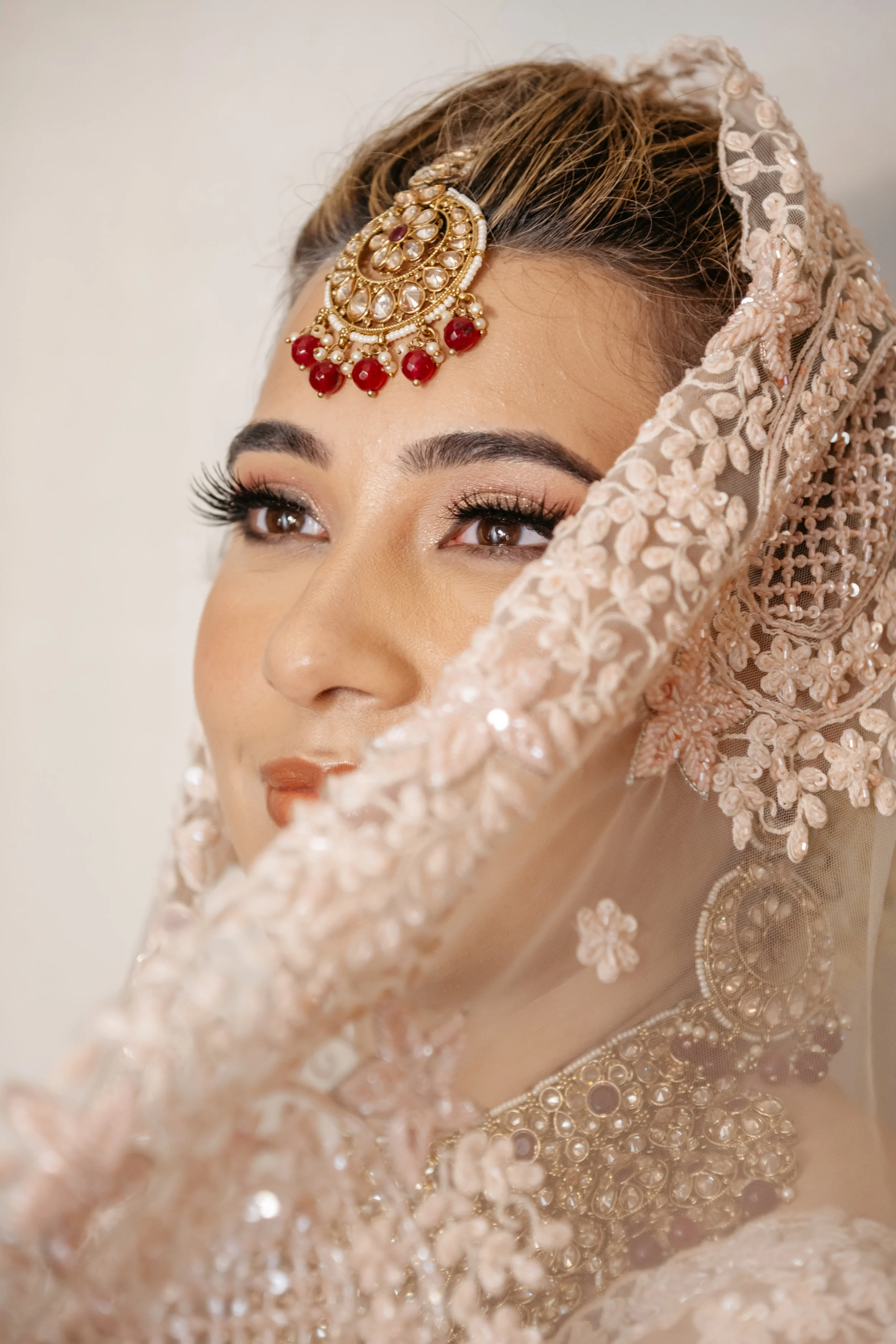 Close-up of a woman wearing elaborate jewelry and a heavily embroidered pink veil, likely a bride preparing for a wedding.