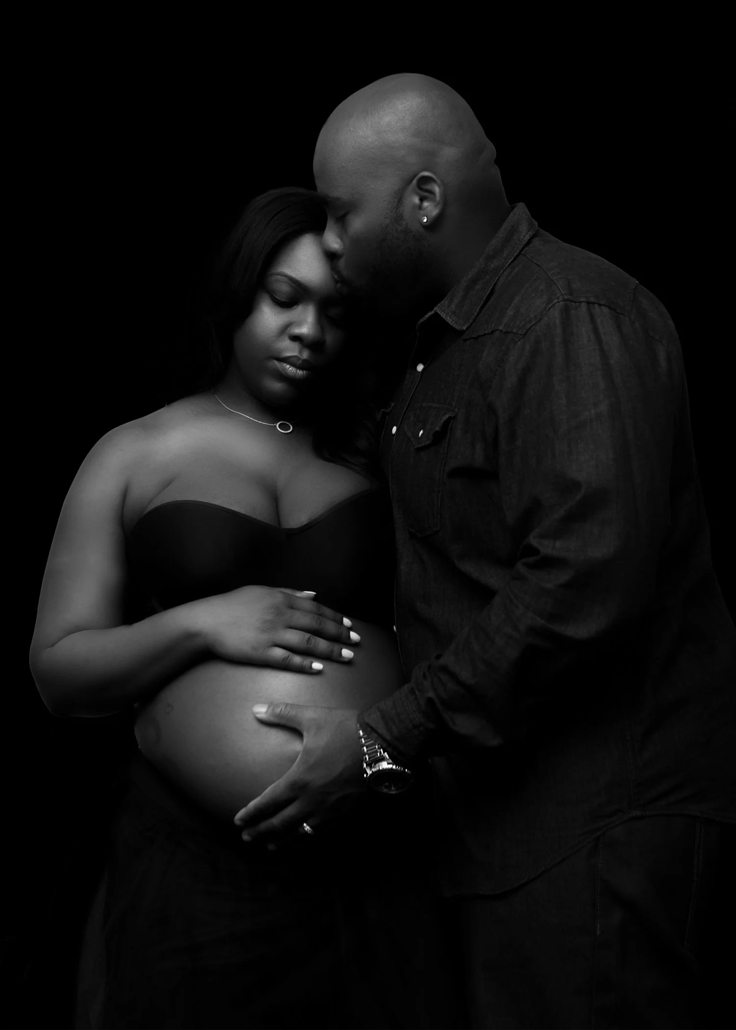 A pregnant woman with dark hair and a strapless top, gently holding her belly, stands next to a man with a shaved head wearing a dark denim shirt. The man is kissing her forehead and cradling her belly with his hand. The scene is in black and white w