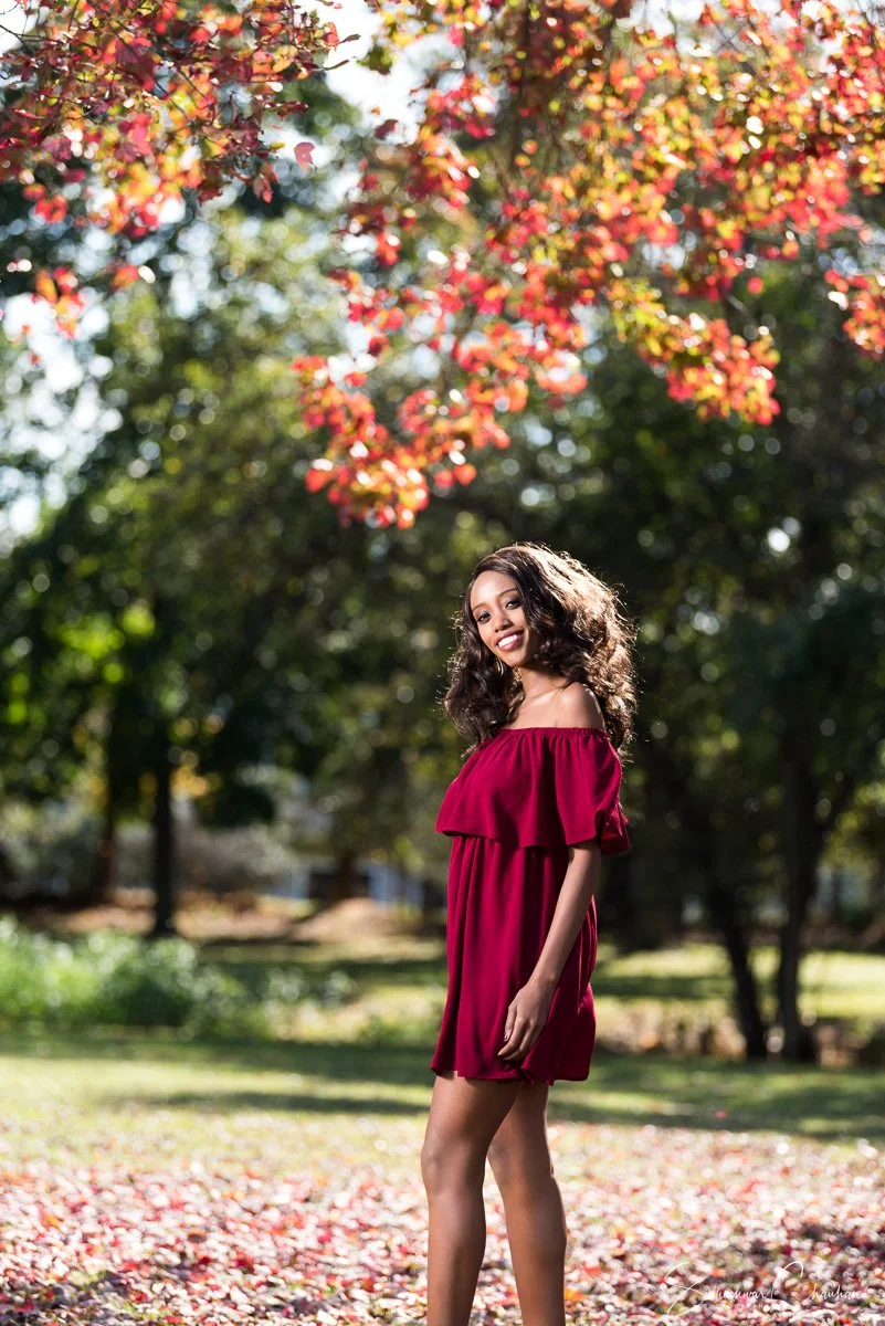 A young woman in a red dress standing in a park with autumn leaves overhead, smiling at the camera.