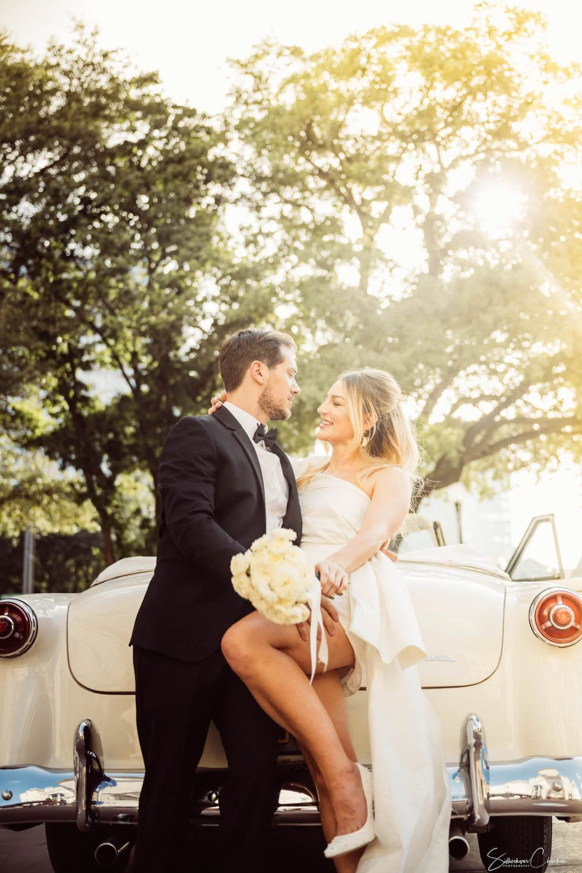 A wedding couple, a bride and groom, standing on the back of a vintage white car outdoors during sunset, with trees and sunlight in the background.