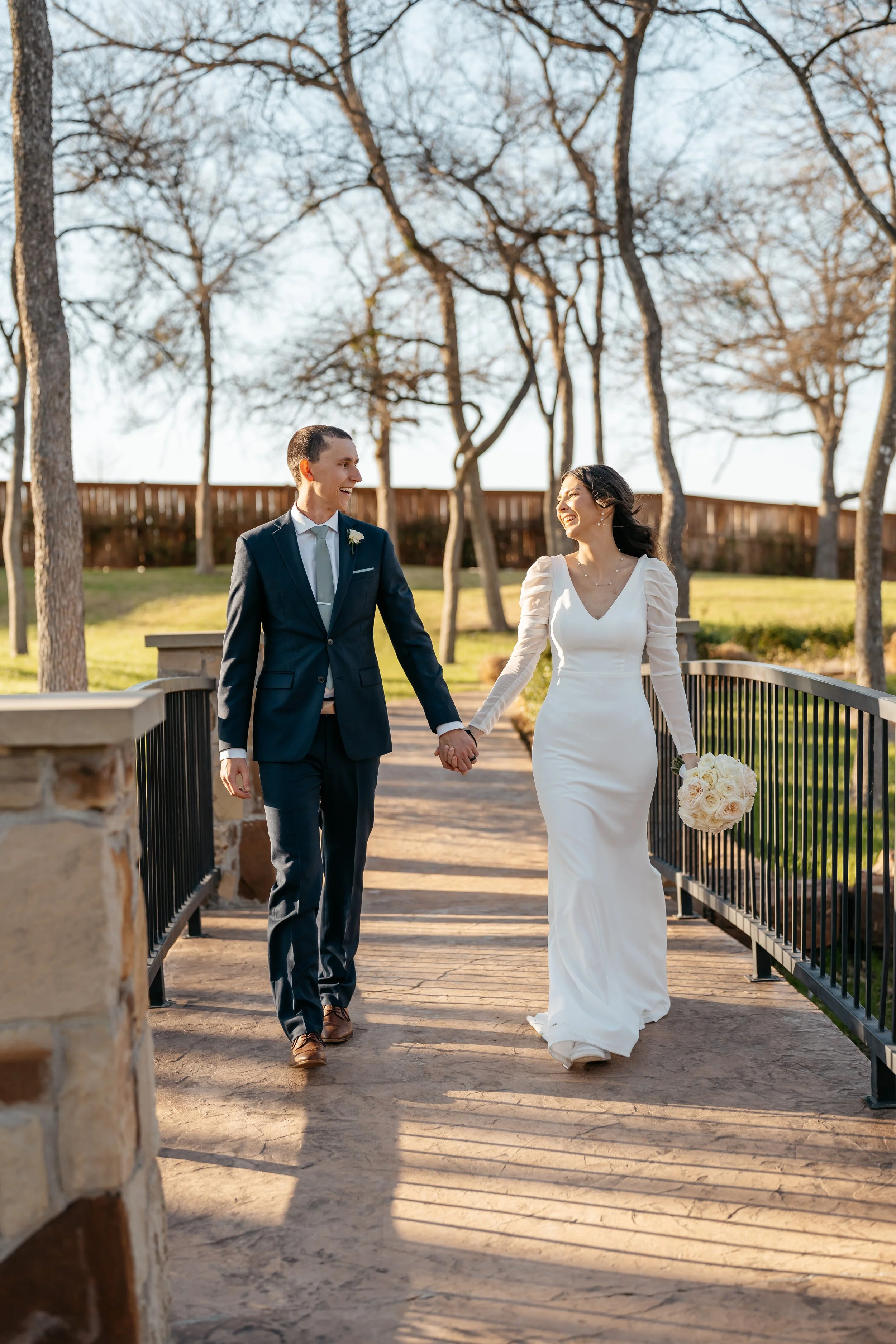 A newlywed couple walking hand in hand on a bridge outdoors, smiling and looking at each other, with trees and a fence in the background.
