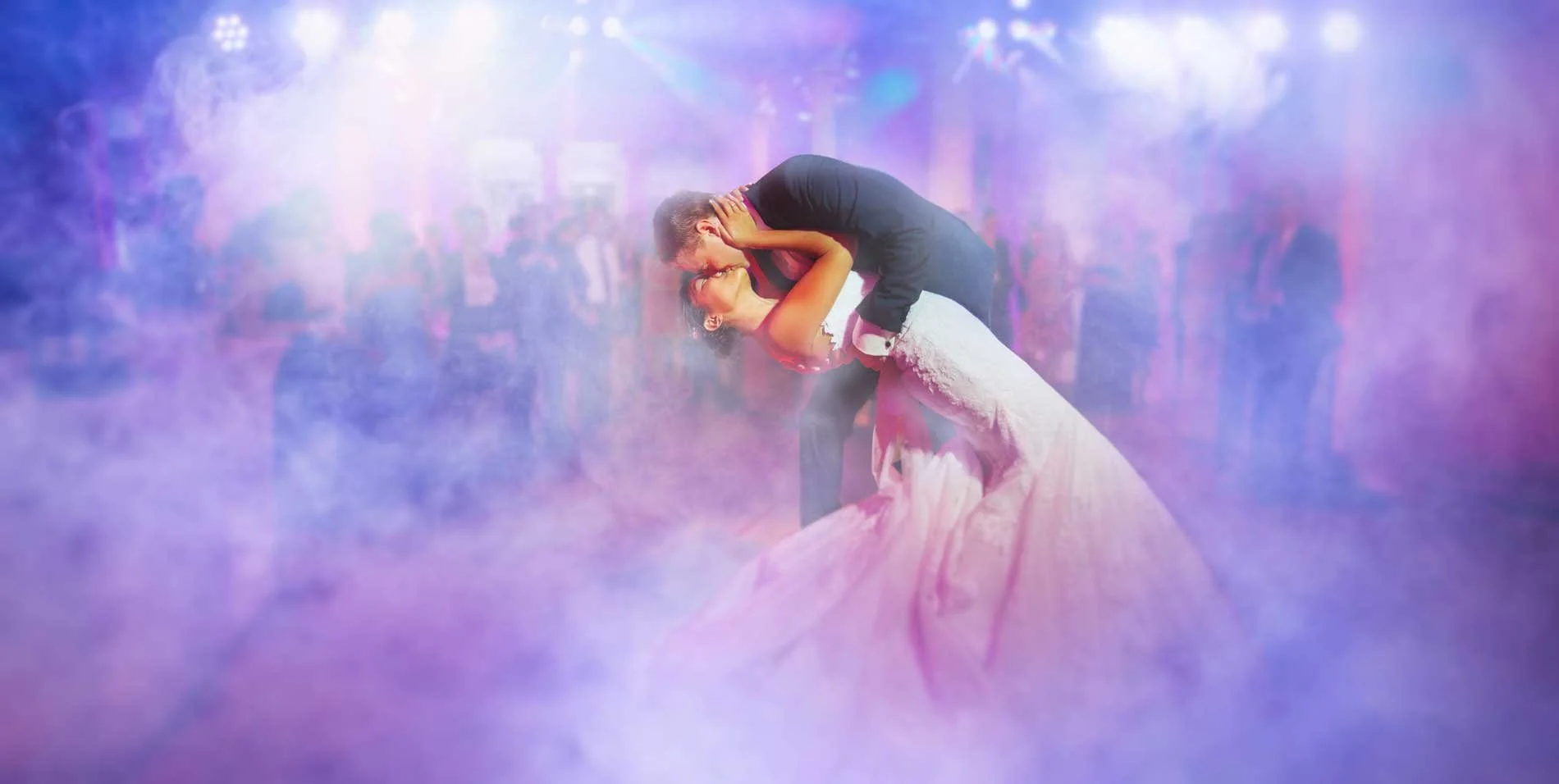 A wedding couple sharing a kiss on the dance floor, with soft pink and purple lighting and a misty atmosphere.