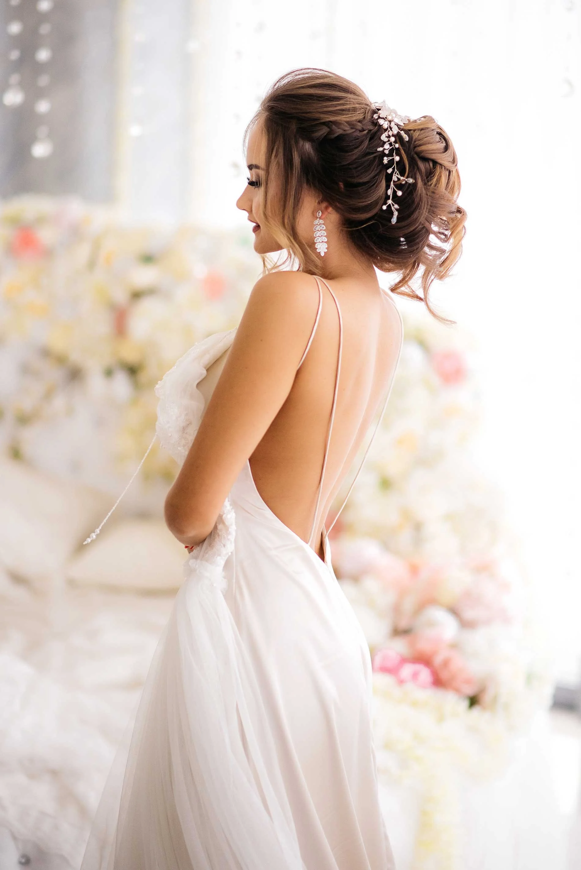 A woman in a white wedding dress with an open back and thin straps, standing sideways, with styled hair adorned with a decorative hairpiece and wearing drop earrings, in front of a floral backdrop.