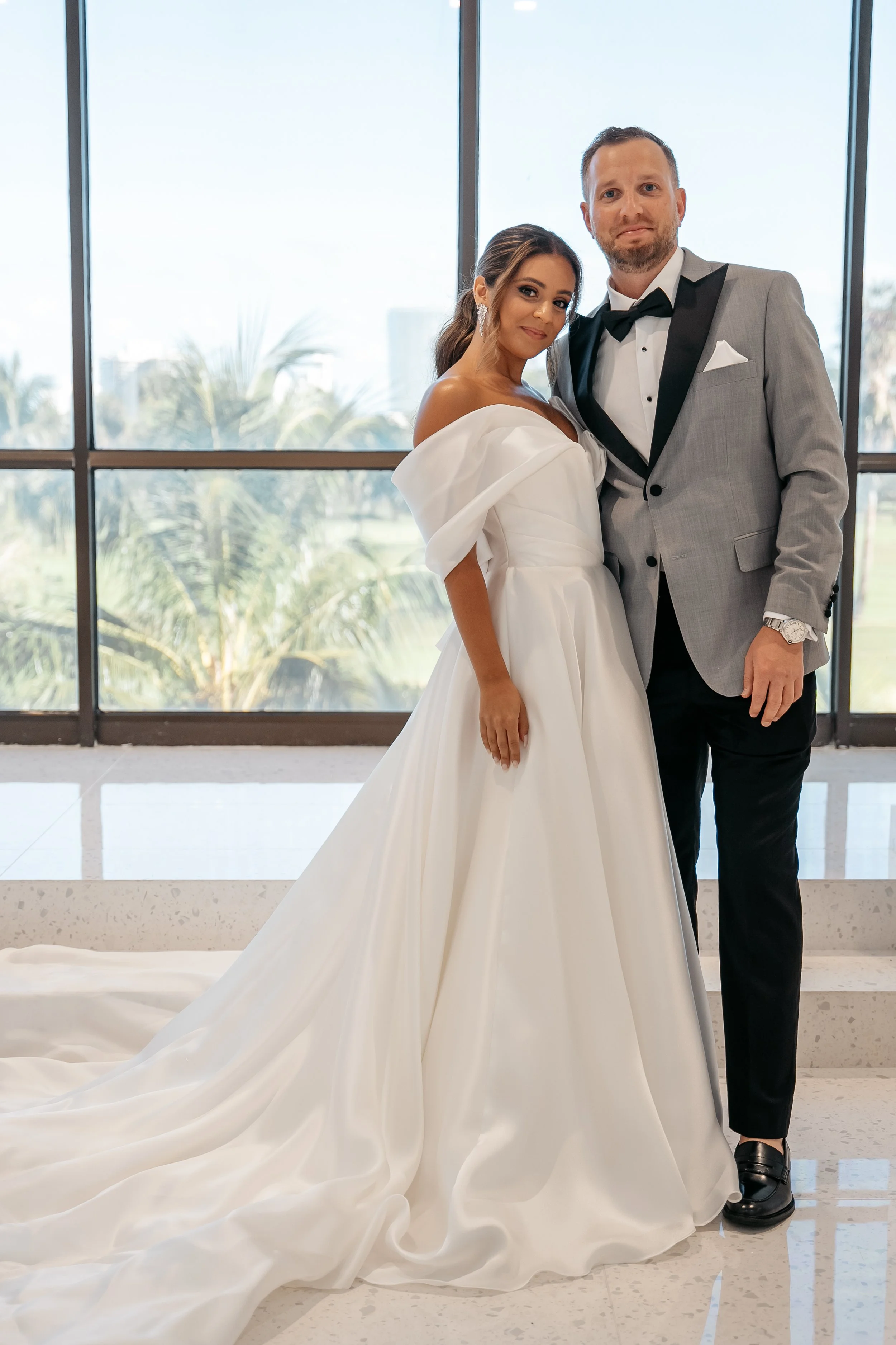 A bride and groom standing together indoors in front of large windows with a view of palm trees, dressed in wedding attire, the bride in a white gown and the groom in a gray tuxedo.