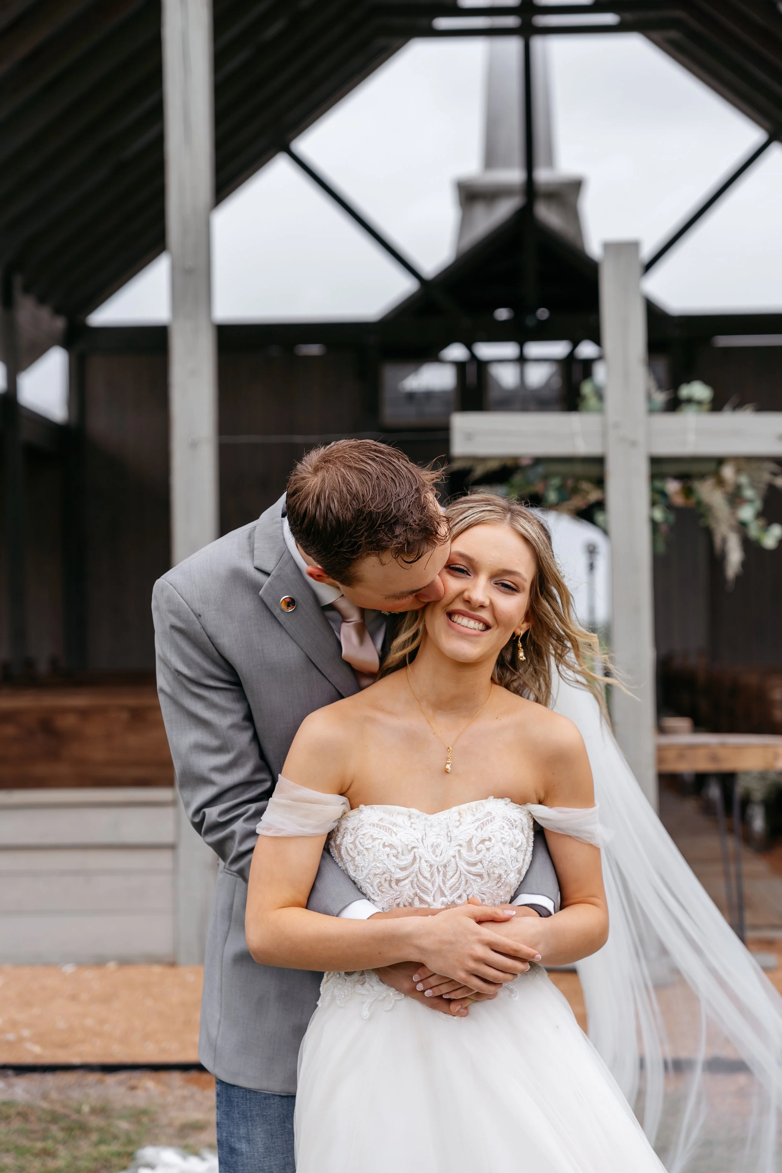 A newlywed couple smiling as the groom kisses the bride on her cheek at an outdoor wedding ceremony.