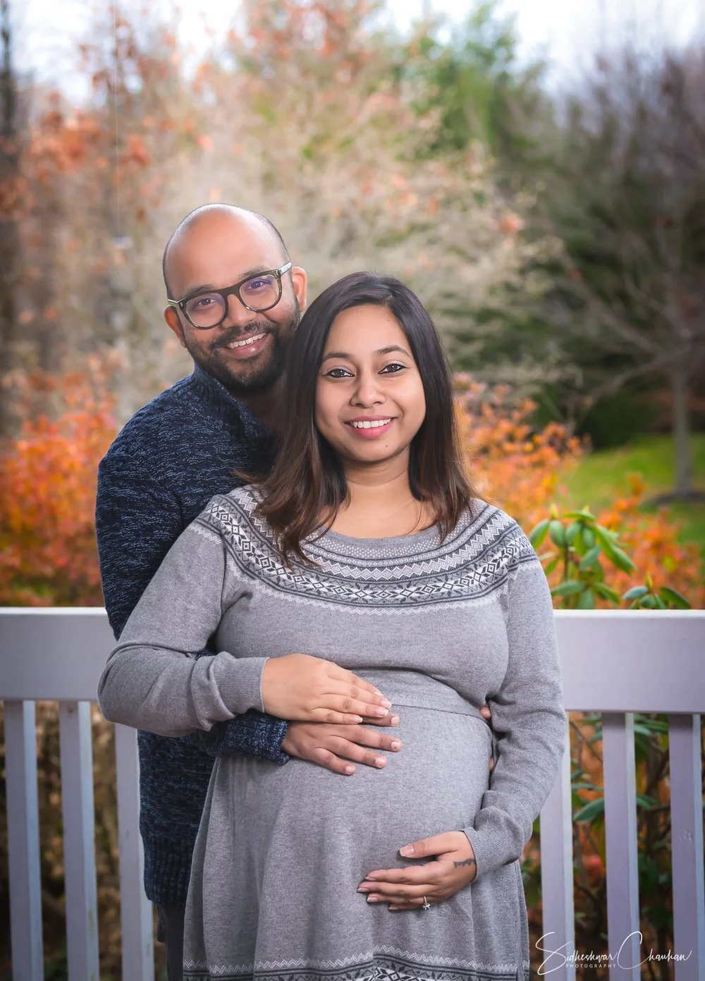 A couple, a man with glasses and a woman with long dark hair, stands outside on a porch railing, with the woman visibly pregnant. They are smiling and facing the camera, with colorful autumn trees in the background.
