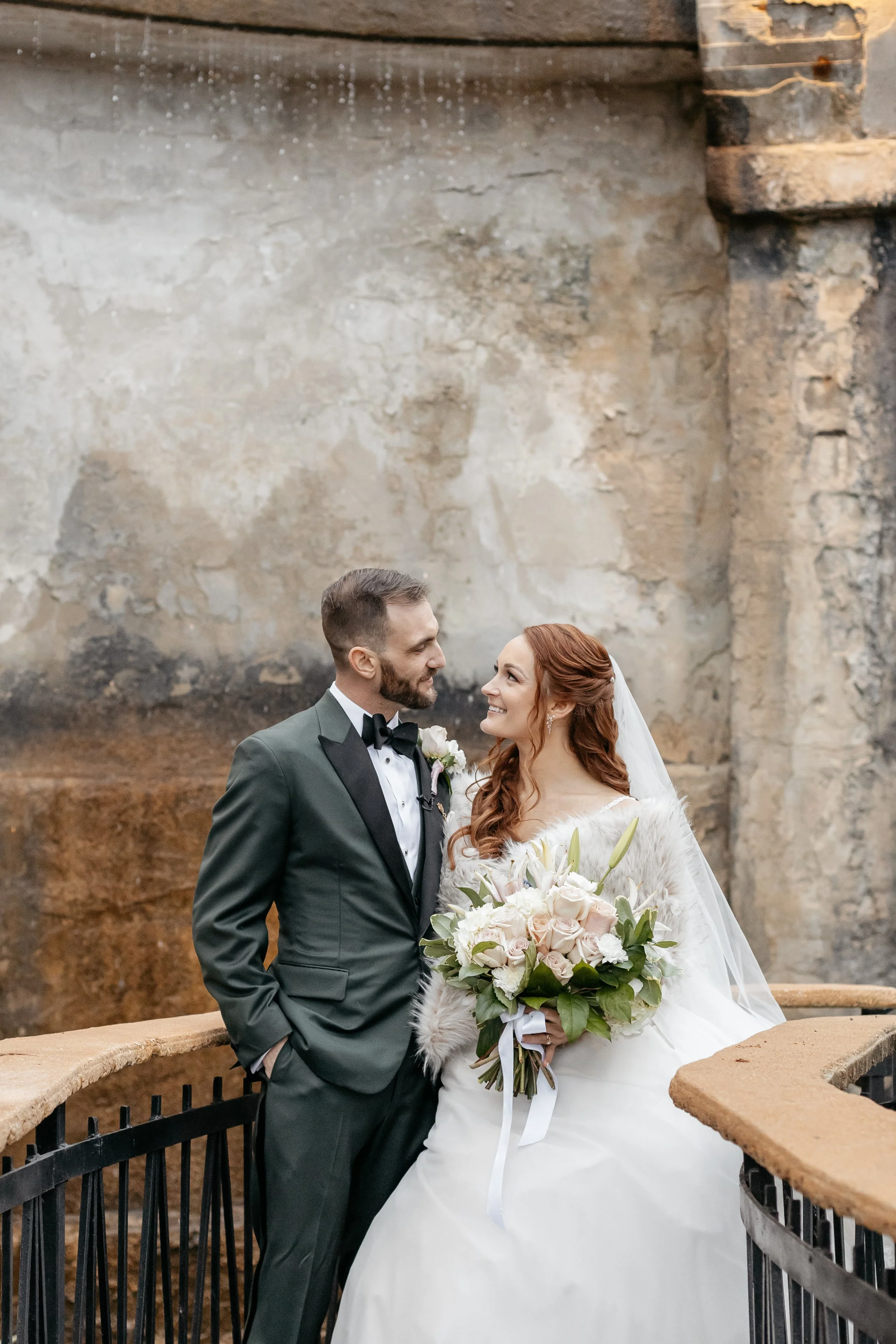 A bride and groom looking at each other, standing on a decorative bridge with stone walls in the background. The bride holds a bouquet of white and blush flowers, and the groom is dressed in a tuxedo.