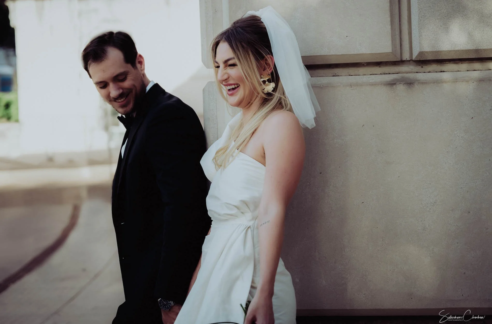 A newlywed couple, a man in a black tuxedo and a woman in a white wedding dress with a veil, leaning against a concrete wall, both smiling and looking down.
