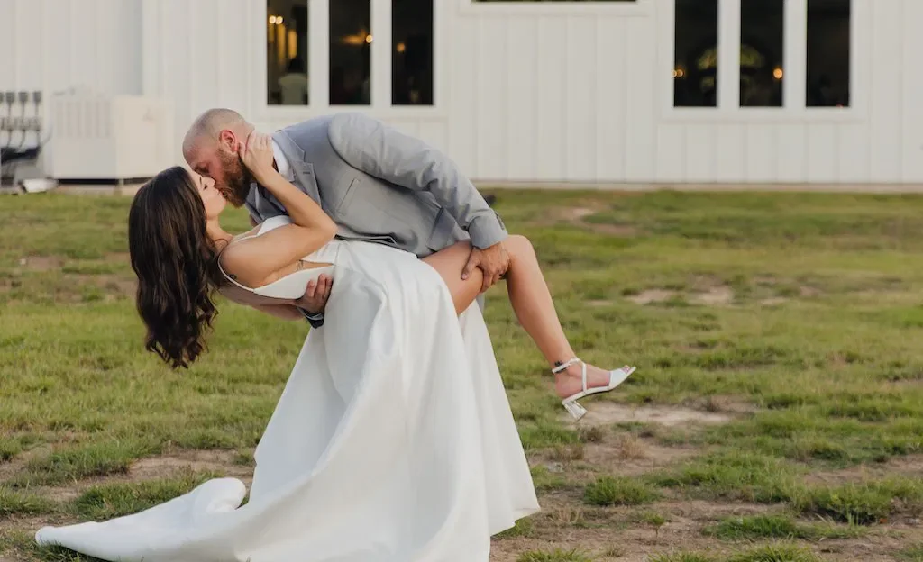 A man in a gray suit dips a woman in a white wedding dress in an outdoor setting with trees and a white fence in the background at sunset.