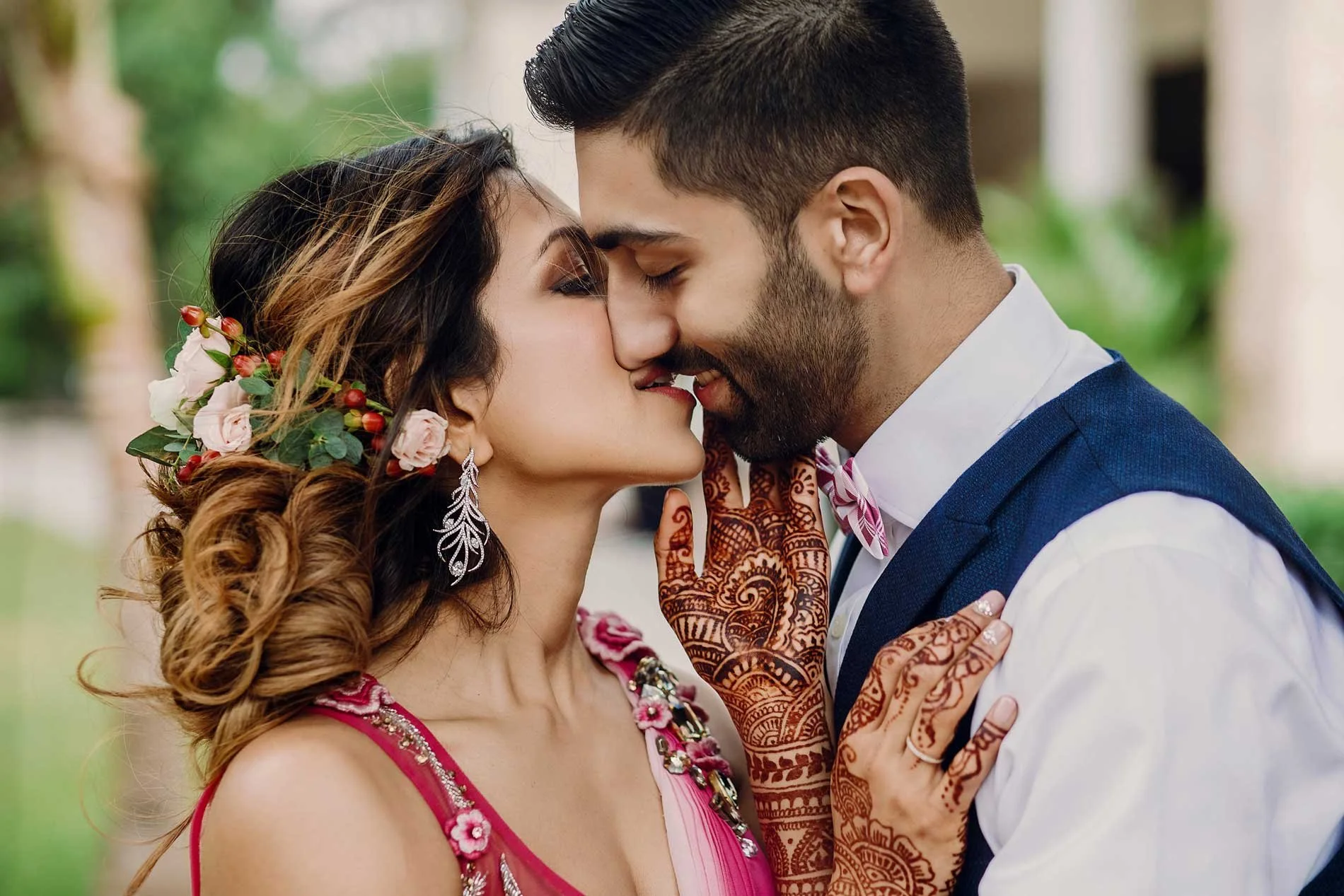 A couple sharing an intimate moment, with their foreheads touching and eyes closed, outdoors with greenery in the background. The woman has floral hair accessories and wears earrings, while the man is in a blue vest with a white shirt and a pink bow