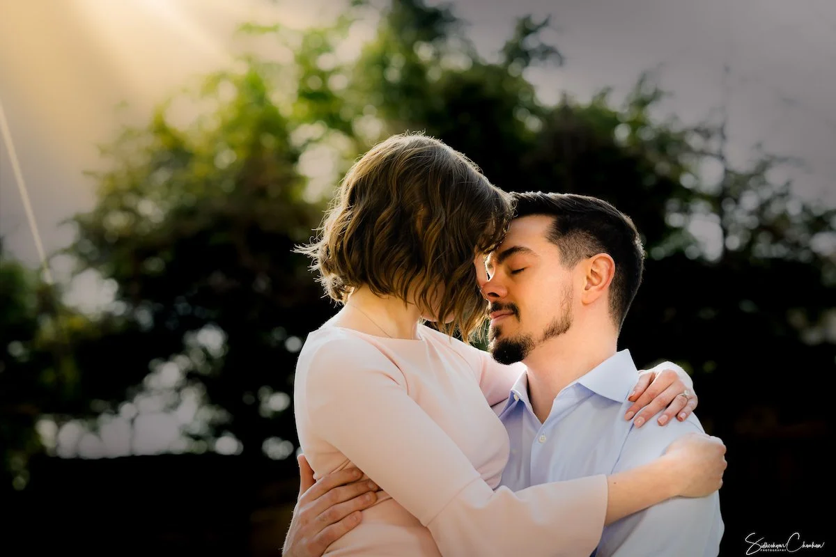 A couple embracing outdoors, with the woman resting her head on the man's forehead, eyes closed, and both with gentle expressions, in front of blurred trees and sunlight.