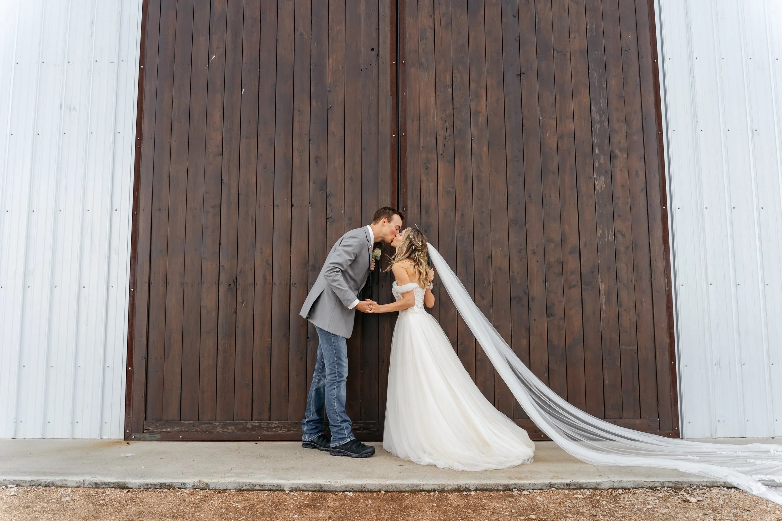 Bride and groom kissing in front of a large wooden barn door, holding hands, with the bride wearing a white gown and long train, and the groom in a gray blazer and jeans.