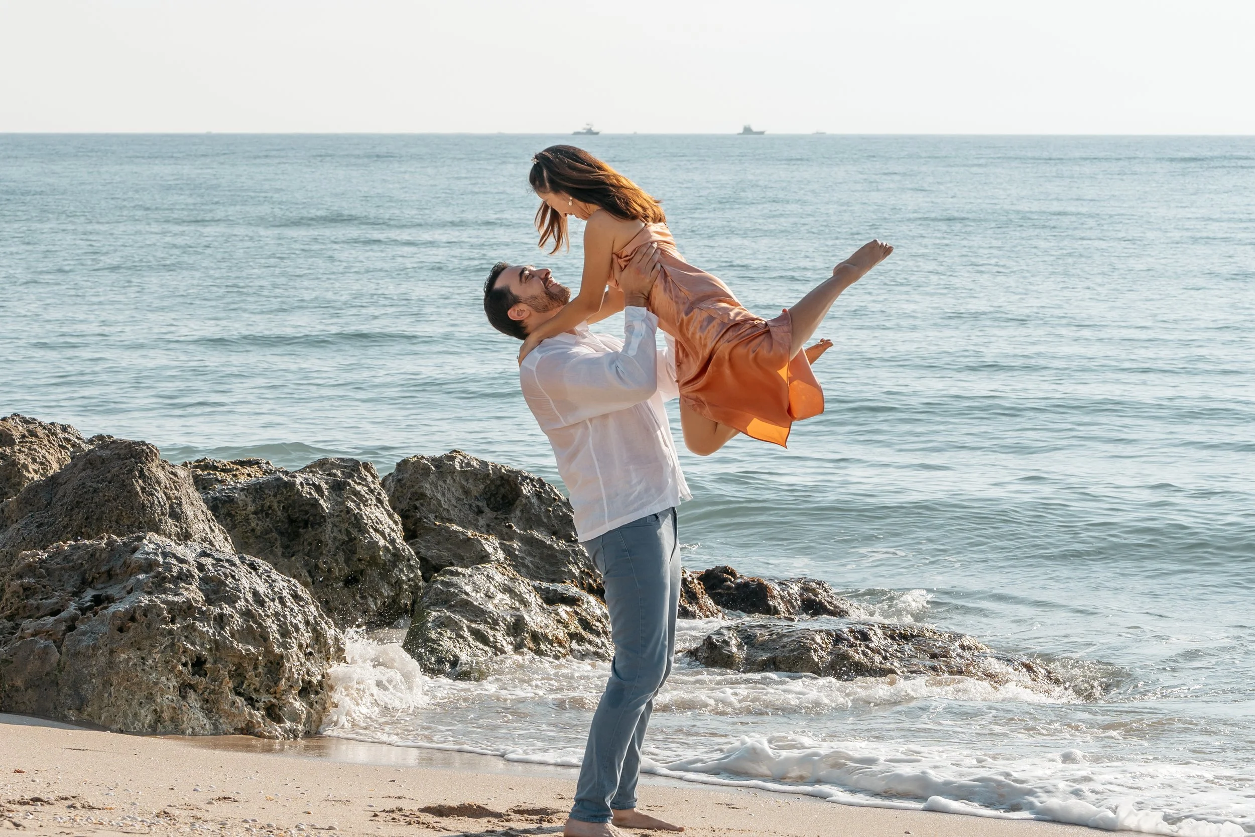 A man lifting a woman in his arms on the beach near rocks, with the ocean and ships in the background.