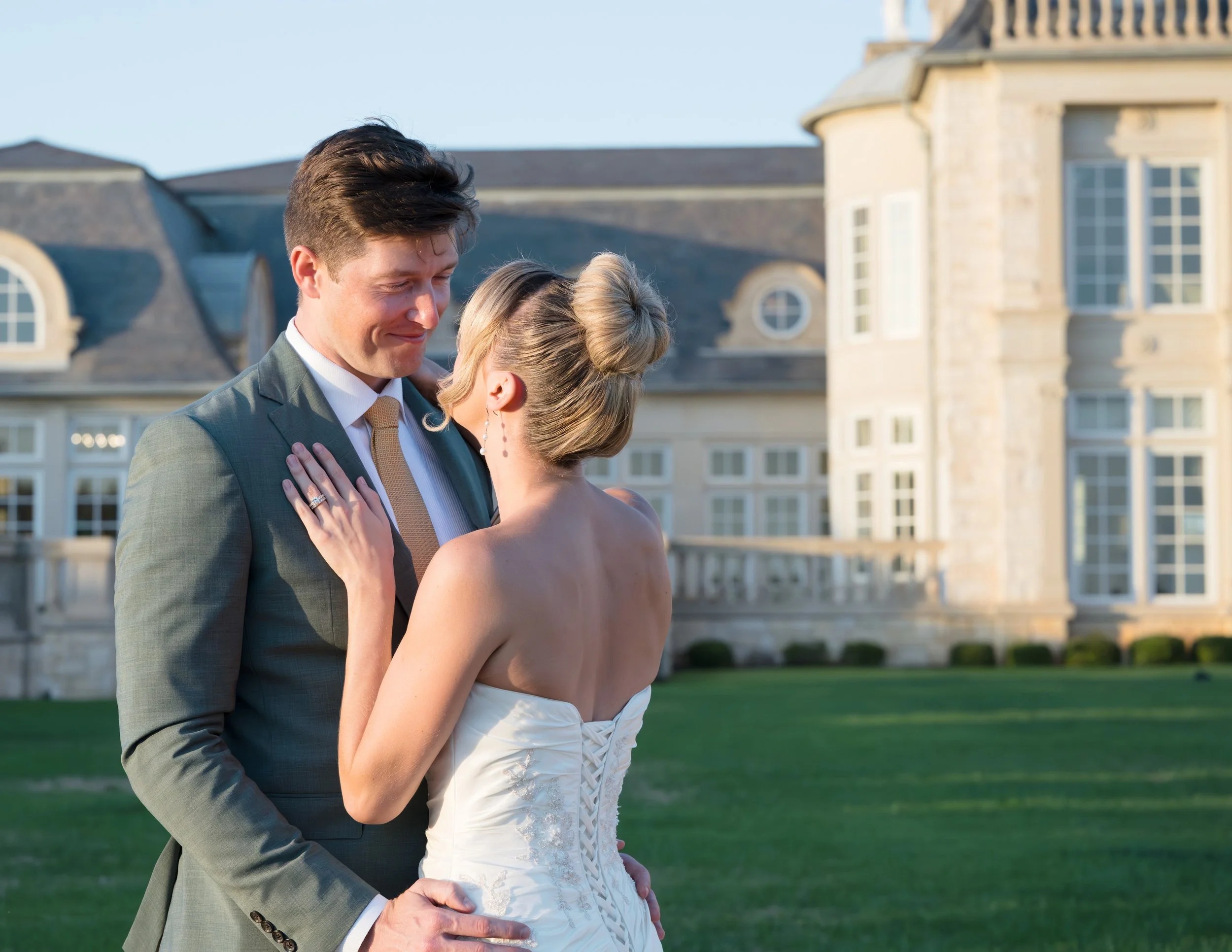 A newlywed couple shares a romantic moment outside a large estate, with the bride in a strapless white wedding dress and the groom in a gray suit, as they look at each other lovingly.