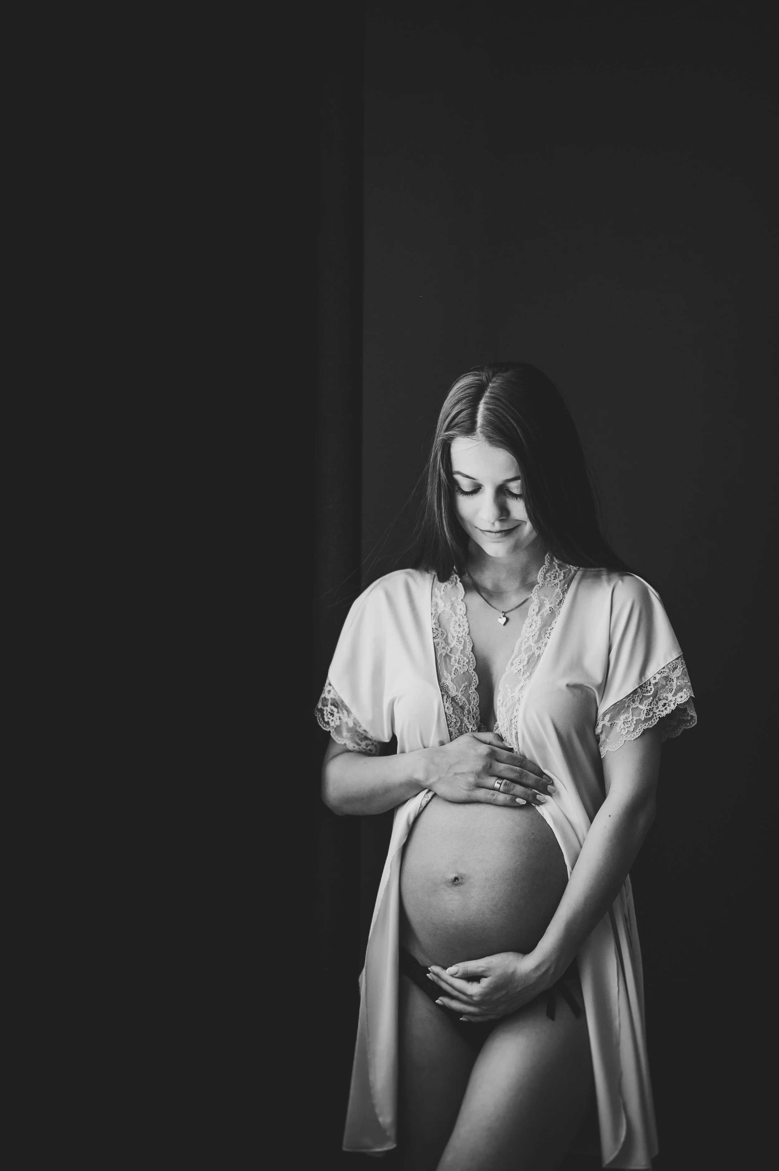 Black and white photo of a pregnant woman in a satin silk robe with lace accents, standing with her hands on her exposed belly, looking down softly.