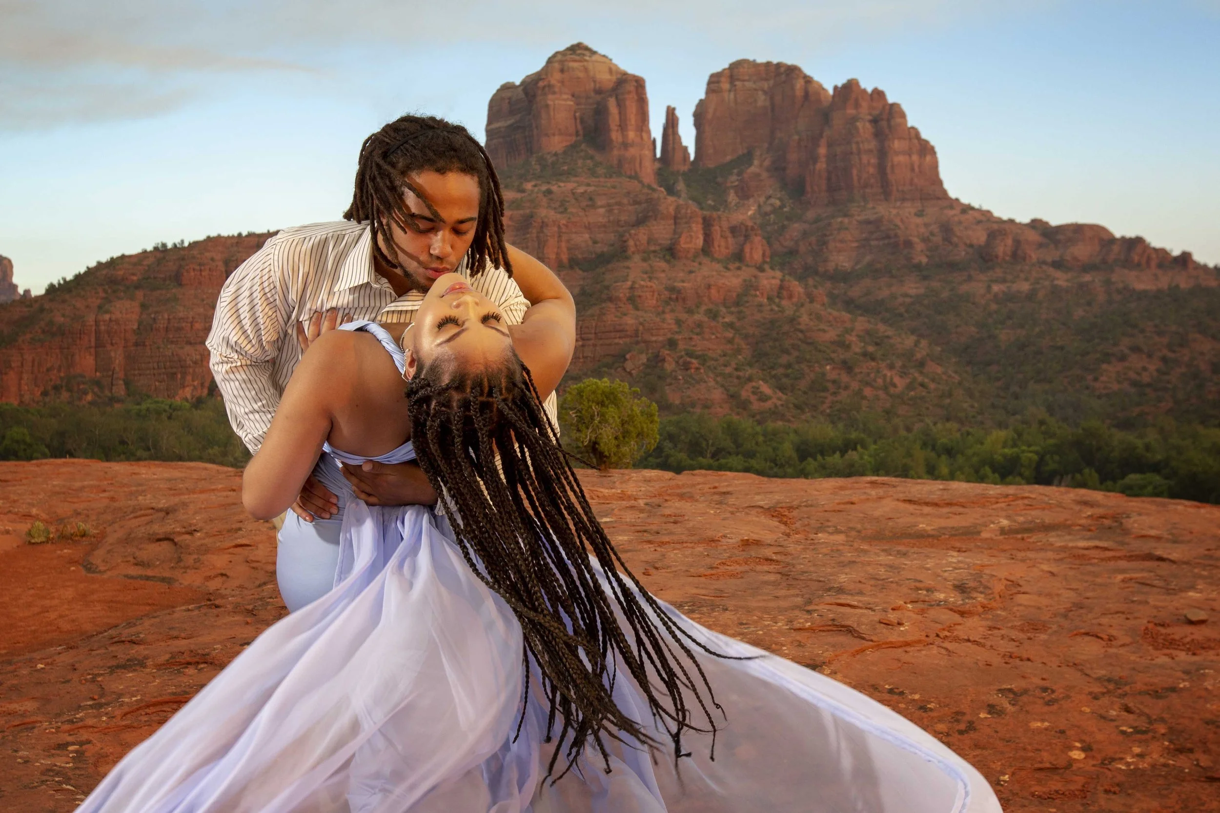 A man with dreadlocks and a woman with braided hair are dancing passionately outdoors on a red rocky landscape with mountains and trees in the background.