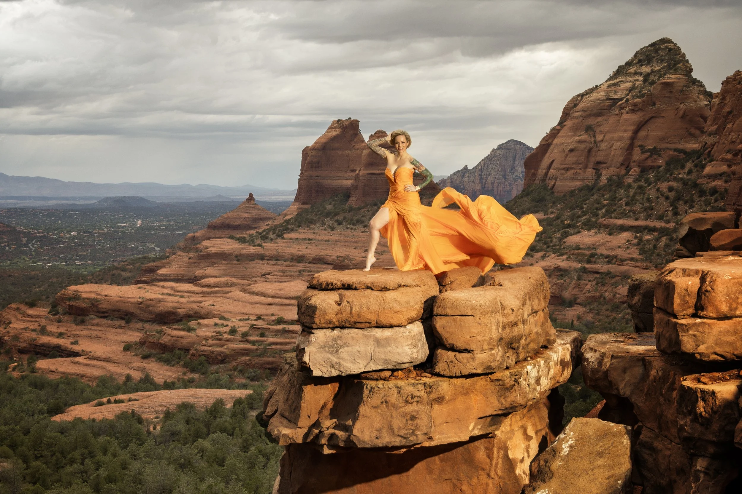A woman in an orange dress standing on a rocky formation in a desert landscape with large red rock formations and a cloudy sky in the background.