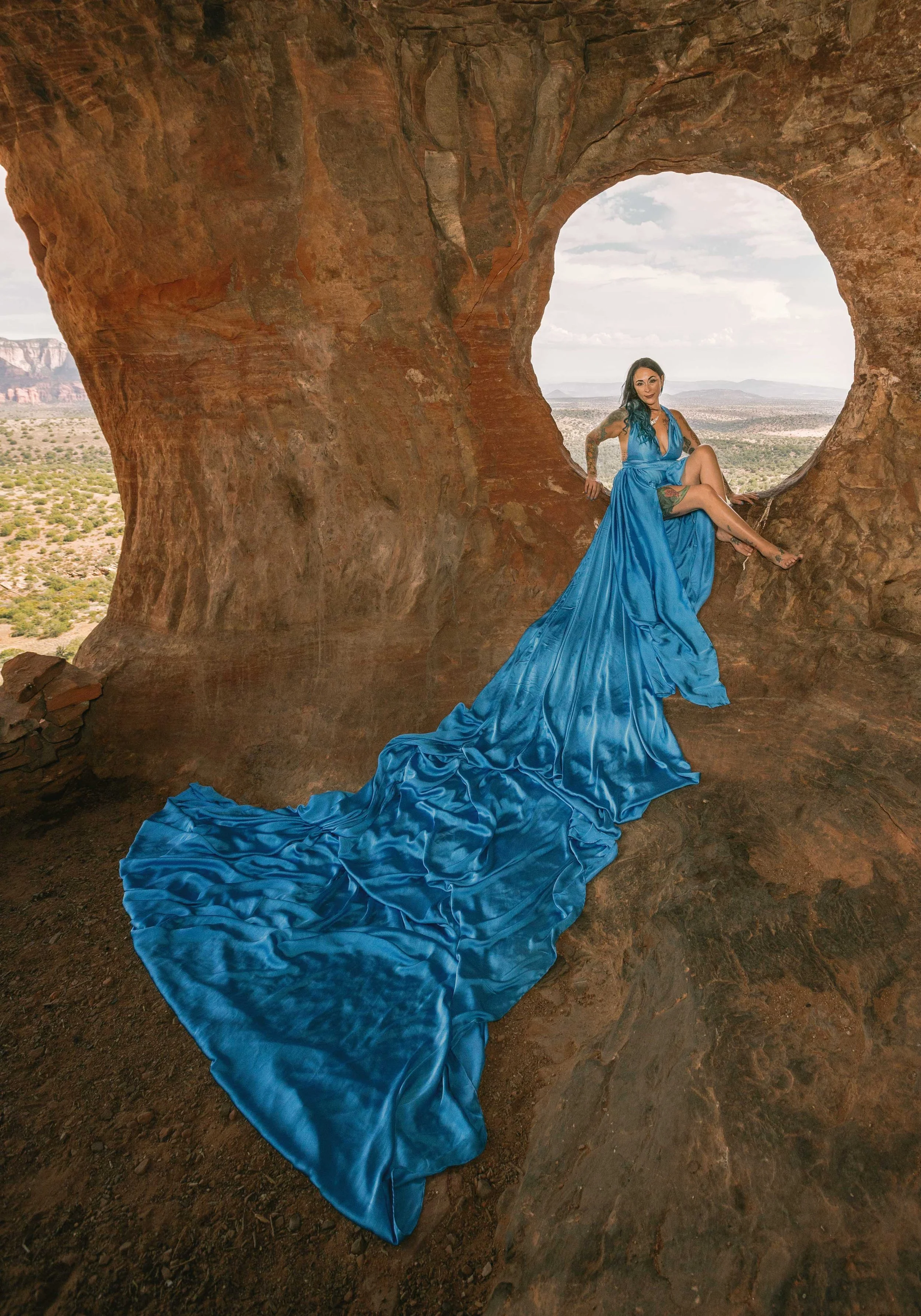 A woman in a long, flowing blue dress sitting inside a natural rock arch in a desert landscape, with sky and distant mountains in the background.