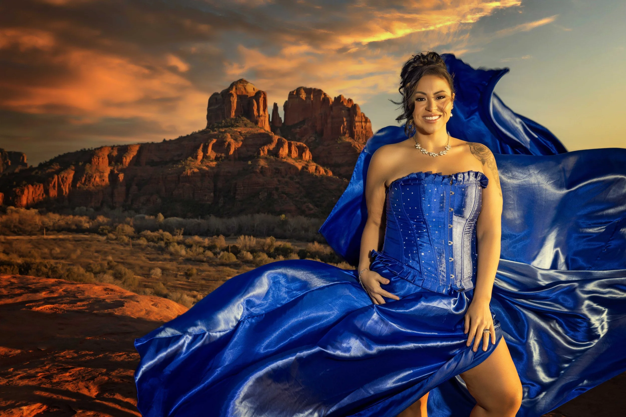 A woman wearing a blue strapless dress and jewelry standing outdoors at sunset with red rock formations in the background.