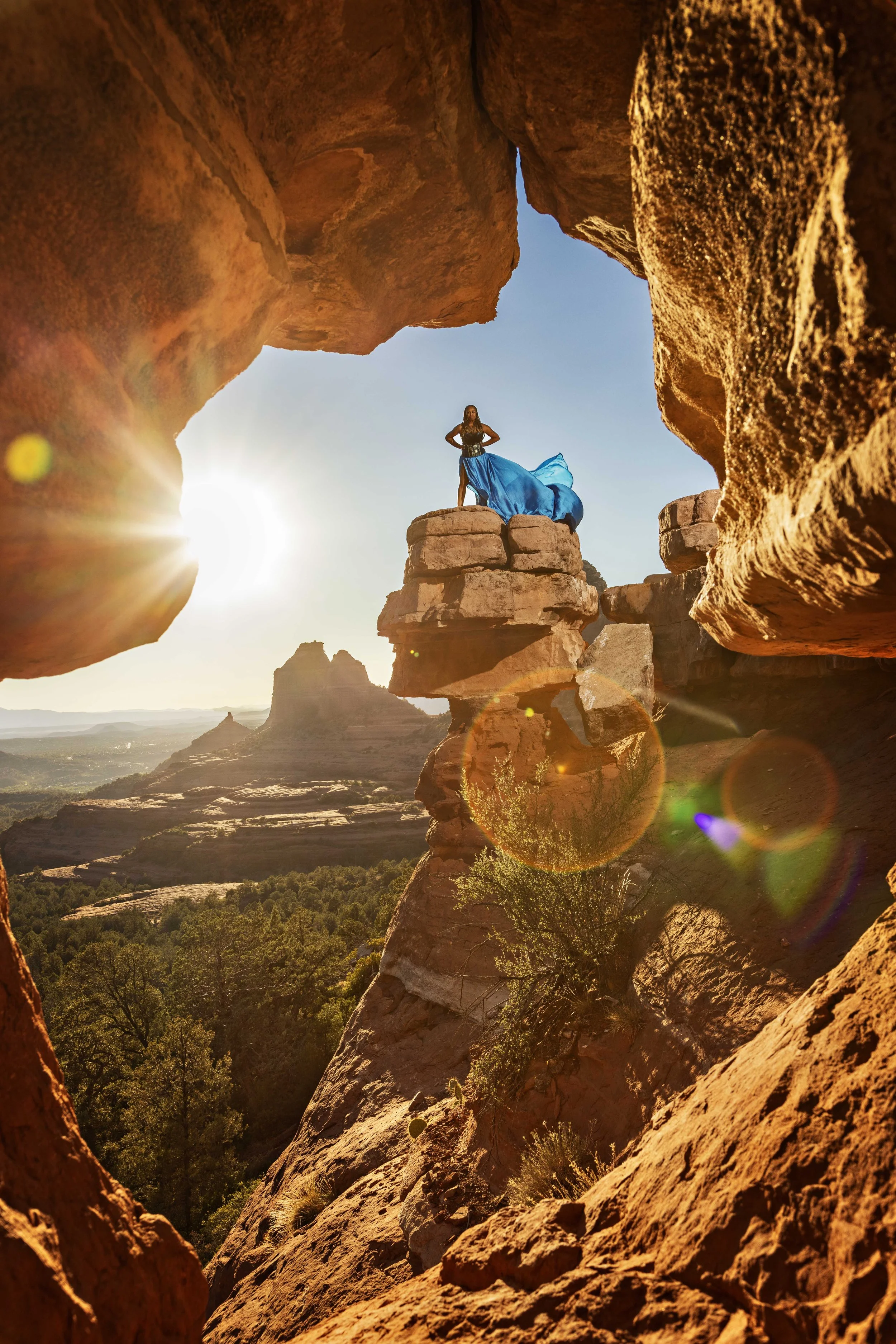 A woman stands on a rock formation in a desert landscape, wearing a flowing blue dress, with the sun setting behind her and creating a lens flare.