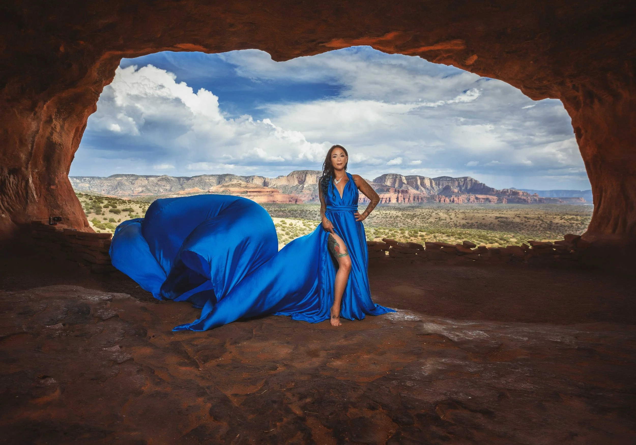 A woman in a long, flowing blue gown with a high slit, standing inside a cave with a view of a desert landscape and red rock formations through the cave opening.