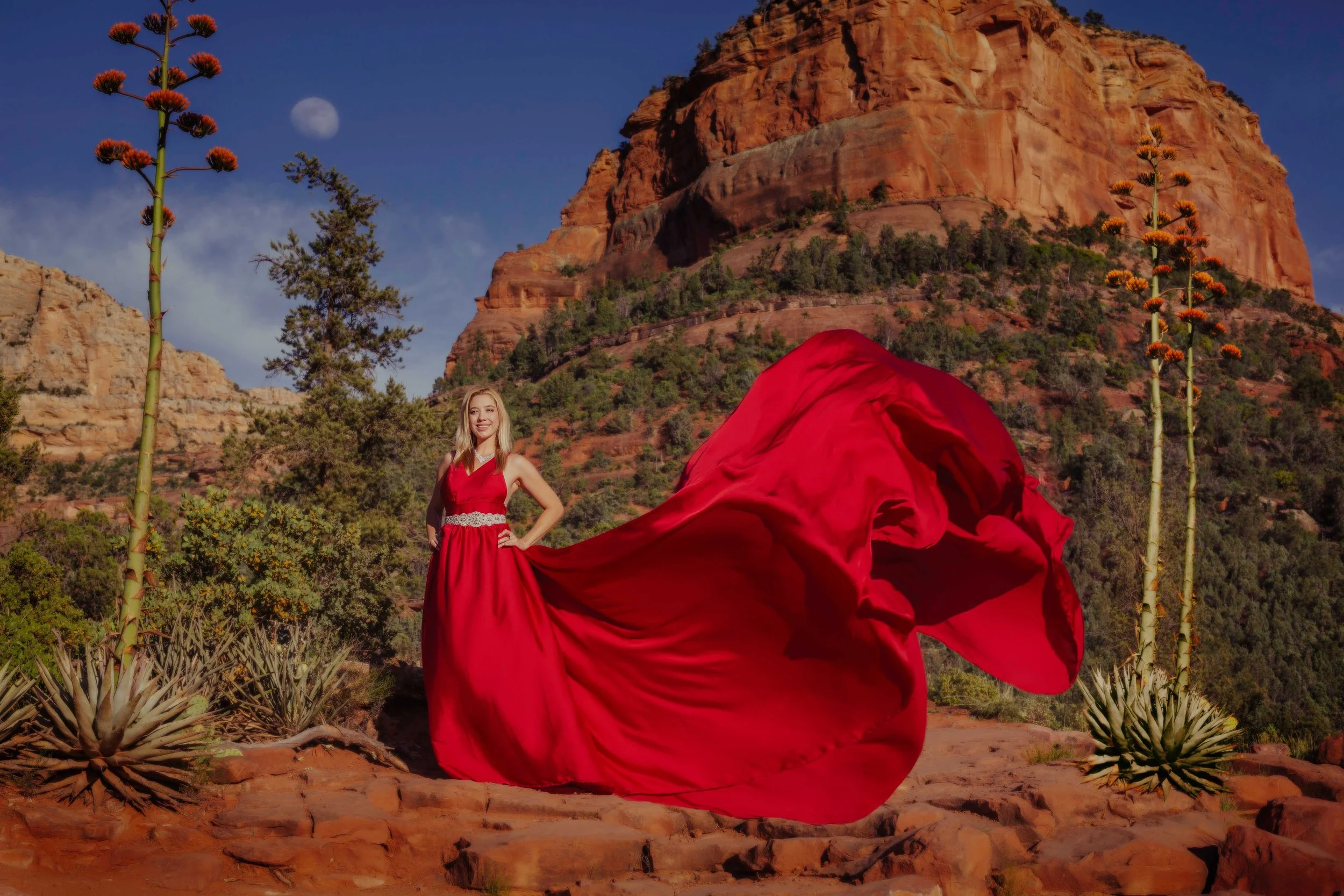 A woman in a flowing red dress standing on a rocky desert landscape with red rock formations and greenery in the background, moon visible in the sky.