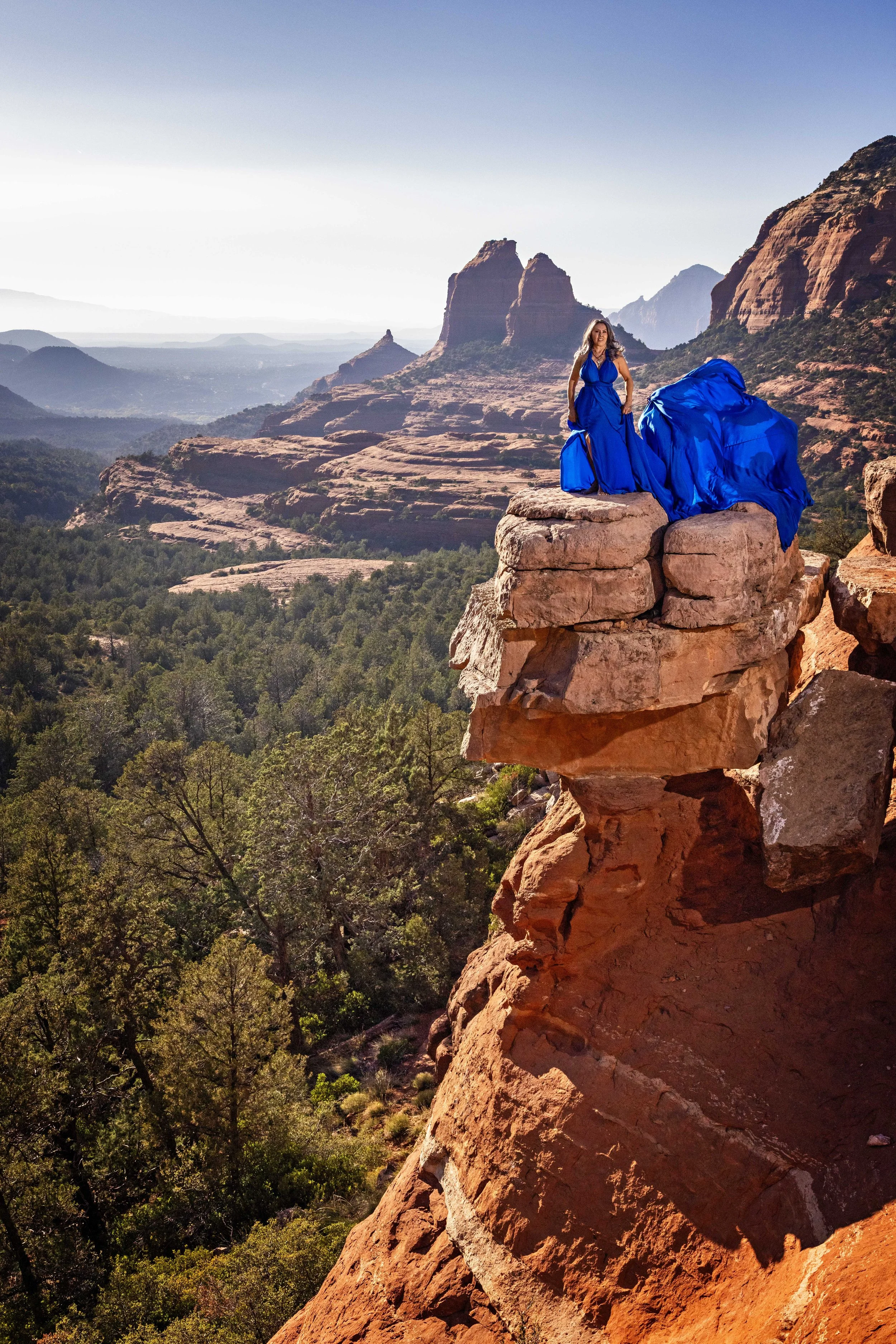 A woman in a blue dress standing on a rock ledge overlooking a desert landscape with red rock formations and green trees, under a clear blue sky.