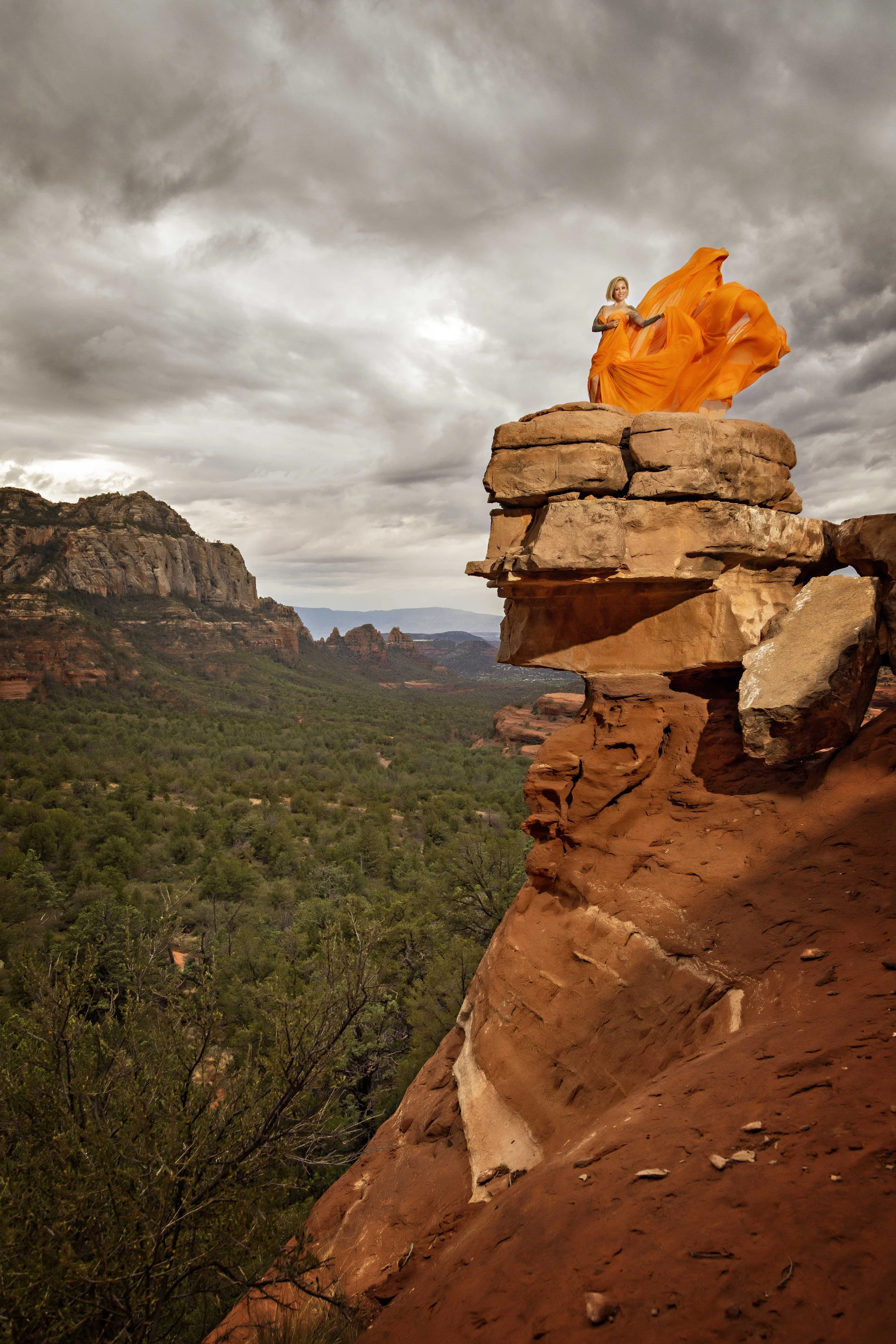 A woman in an orange dress standing on a rocky ledge overlooking a desert landscape with green trees and distant red rock formations under a cloudy sky.