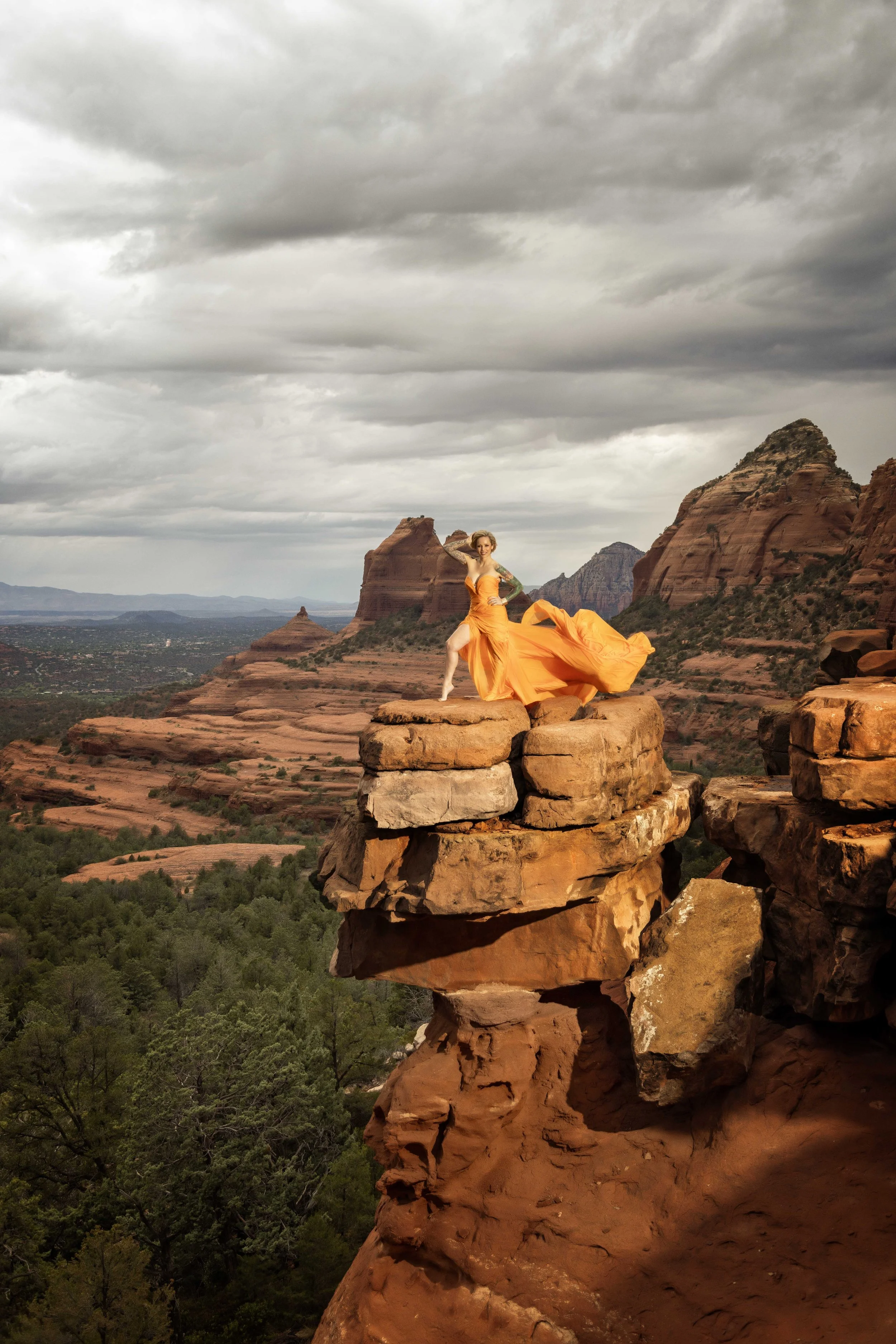 A woman in an orange dress posing on a rock formation in a desert landscape with mountains and cloudy sky in the background.