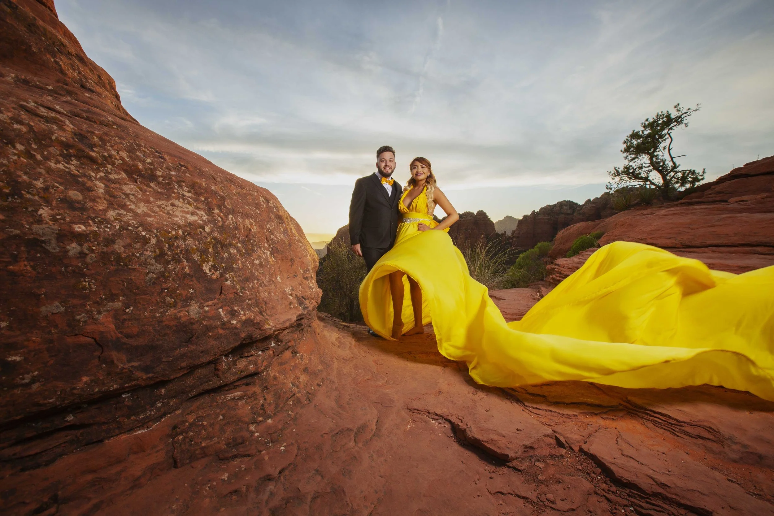 Couple standing in a desert landscape at sunset, with large red rocks and sparse vegetation. The woman is wearing a long, flowing yellow gown, and the man is dressed in a black suit with a yellow bow tie.