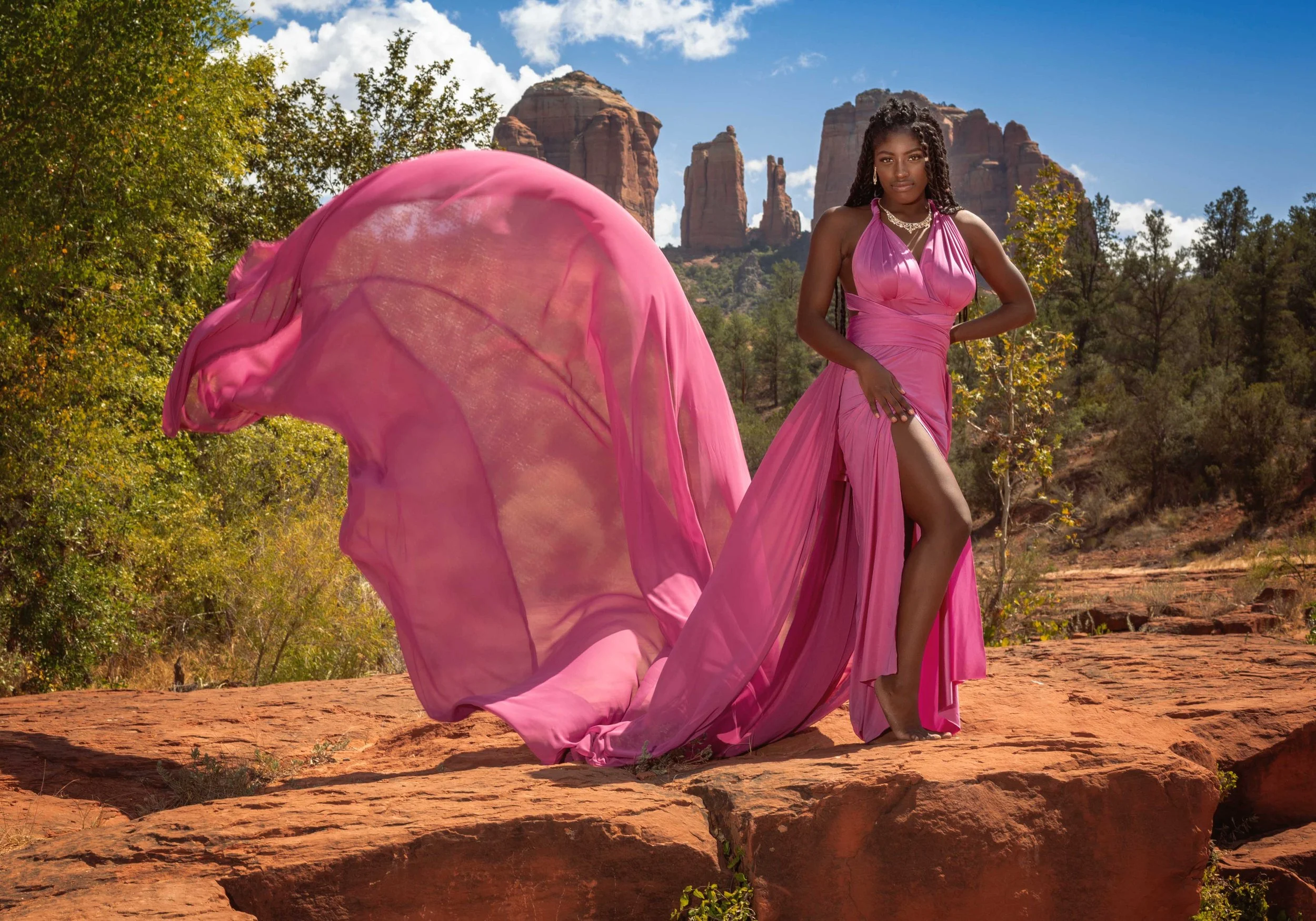A woman in a pink flowing gown with a slit stands on red rocky terrain with red rock formations and green trees in the background.