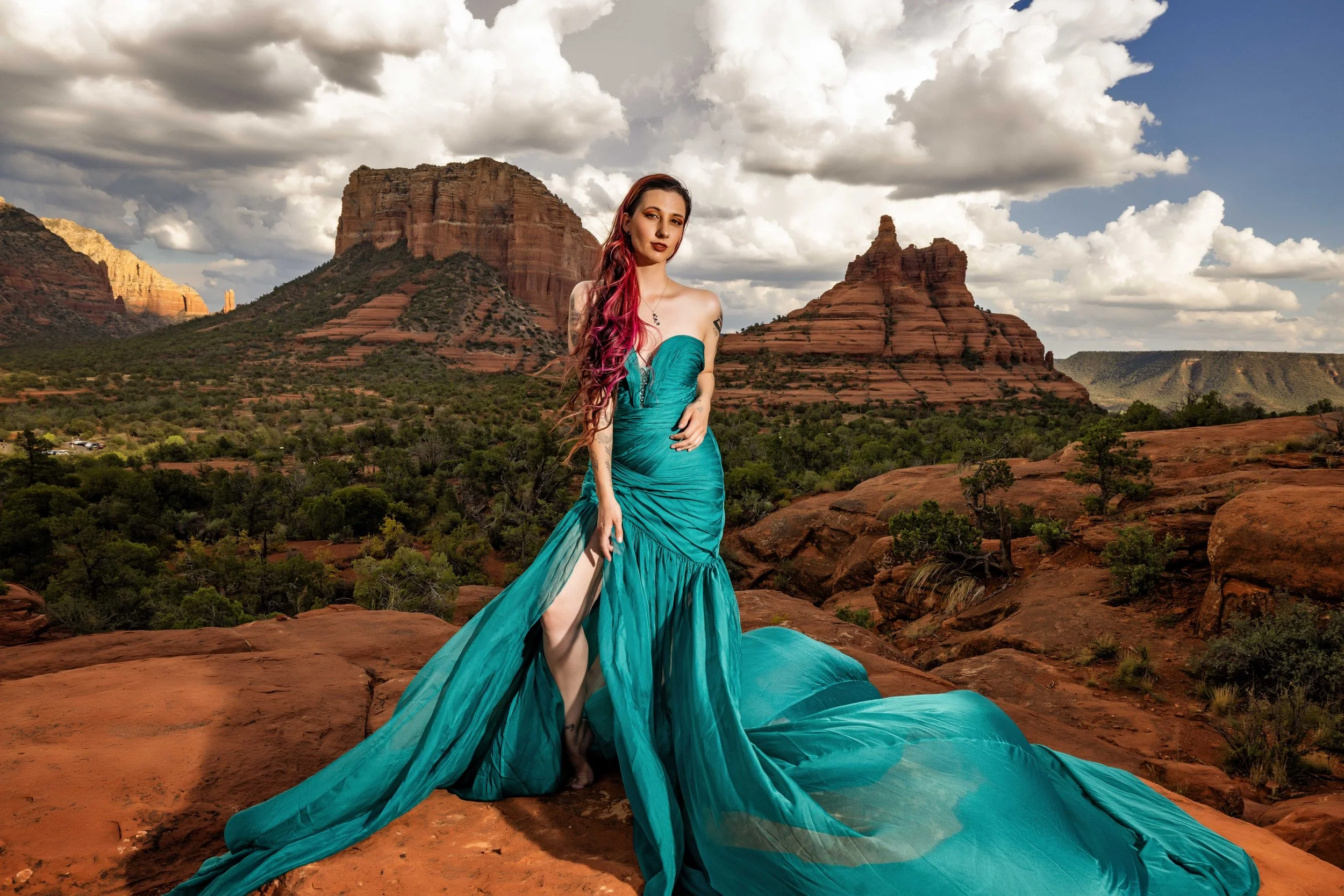 A woman in a flowing turquoise dress stands on red rock terrain with desert vegetation, with large red rock formations and a partly cloudy sky in the background.