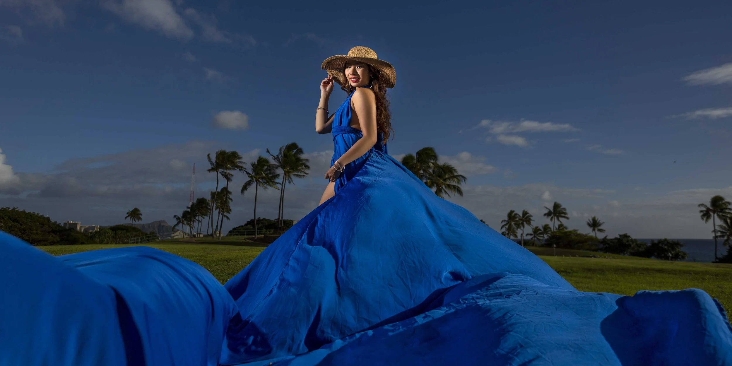 lady in a blue flying dress wearing a big hat smiling with palm tress in the background