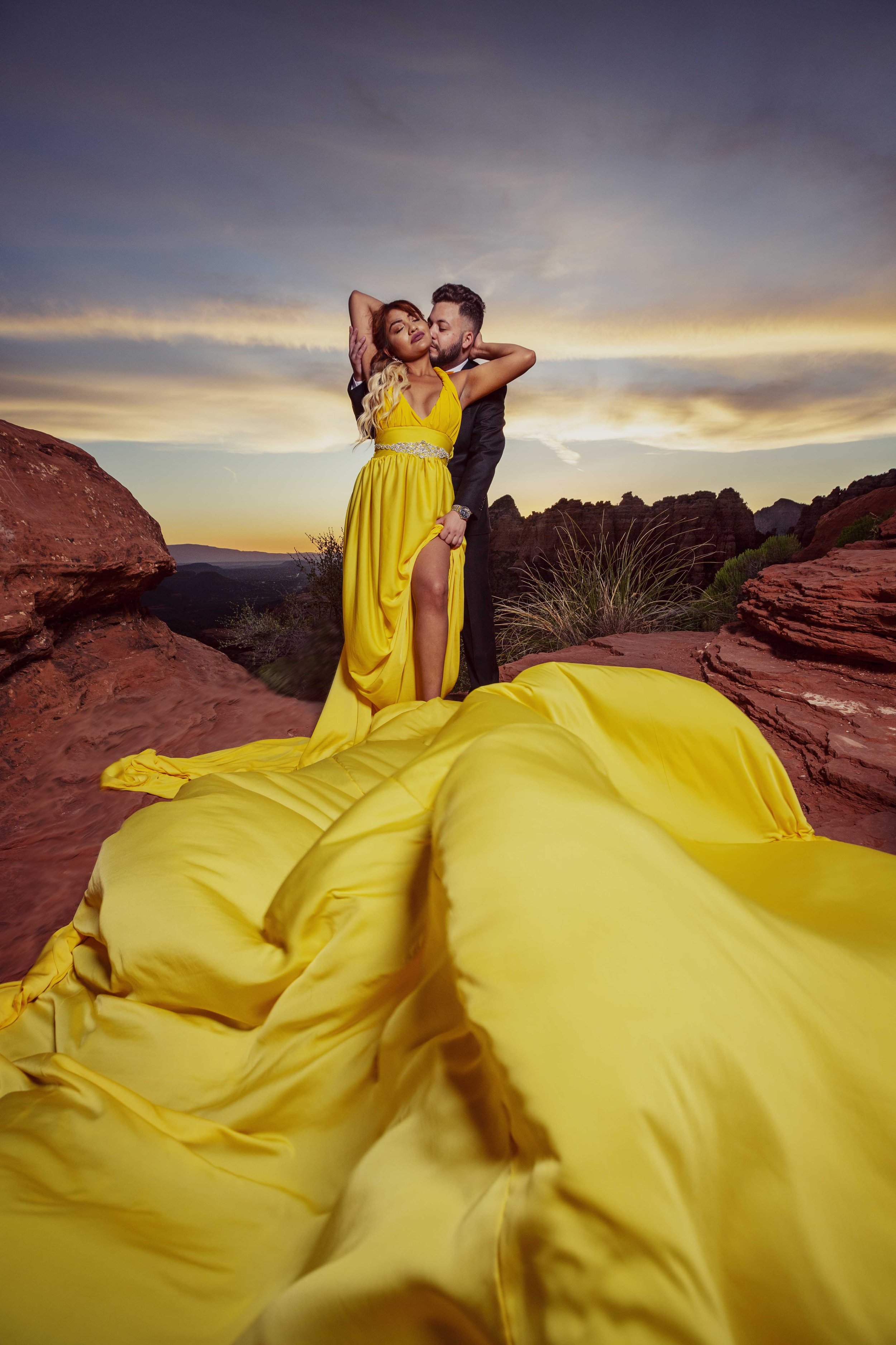 A romantic couple dancing outdoors on a rocky landscape during sunset, with the woman in a vibrant yellow gown and the man in a black suit, and a flowing yellow train on the ground.