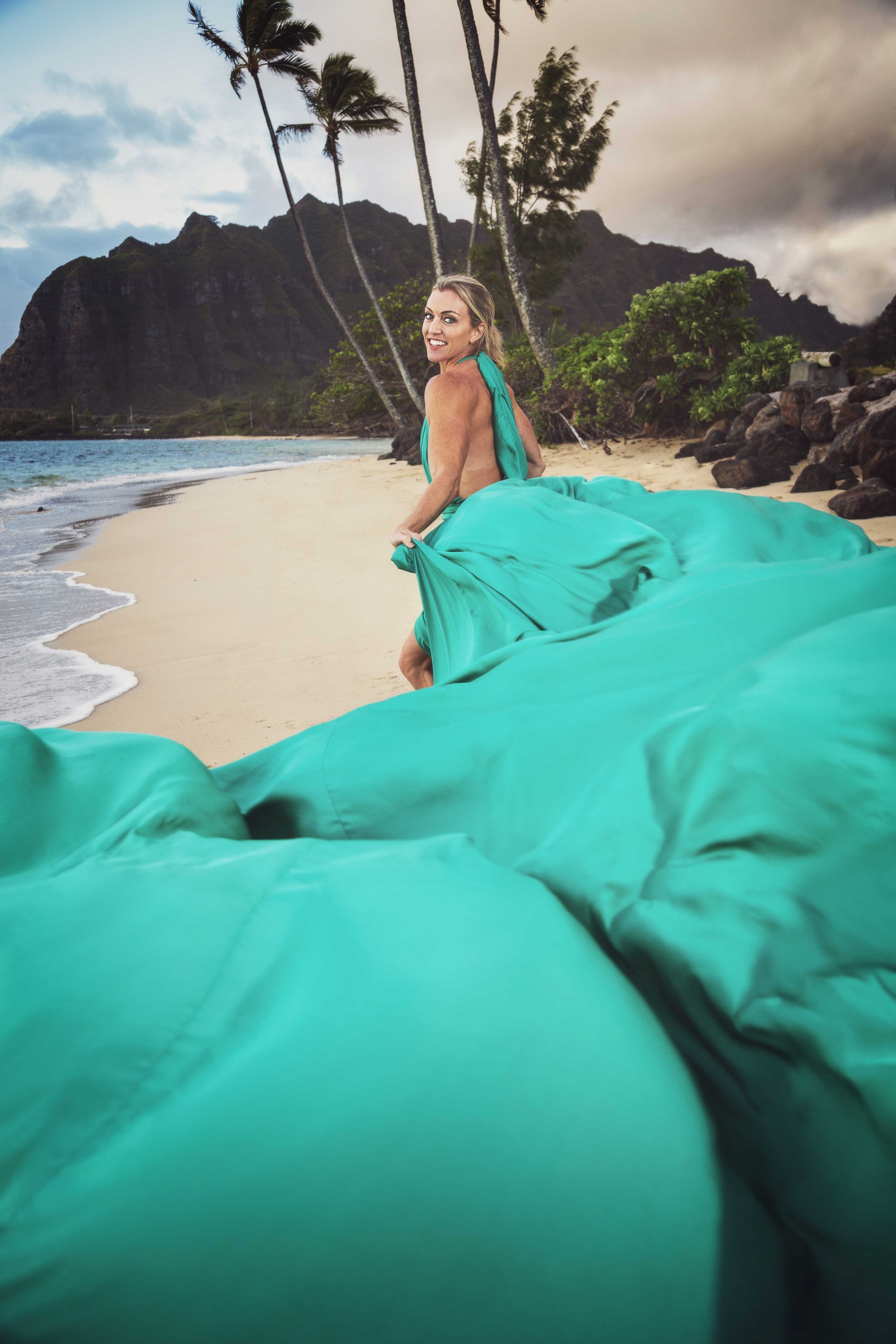 Woman in a turquoise dress on a sandy beach with palm trees, rocky shoreline, mountains, and stormy sky in the background.