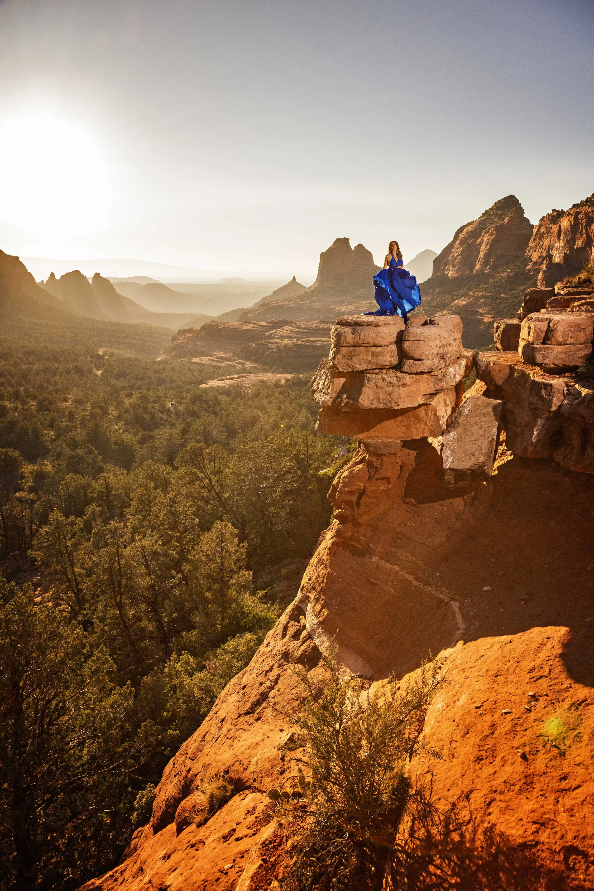 A woman in a blue dress standing on a rocky ledge overlooking a canyon during sunset.