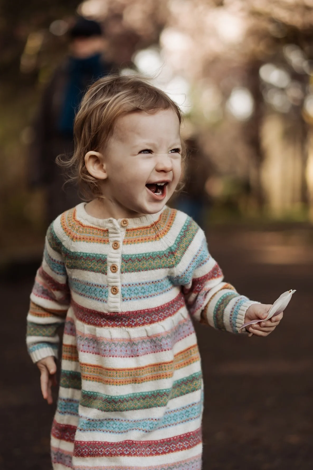 A young girl with curly red hair is smiling and laughing outdoors, wearing a colorful striped knit dress. In the background, a person with a backpack is slightly blurred.